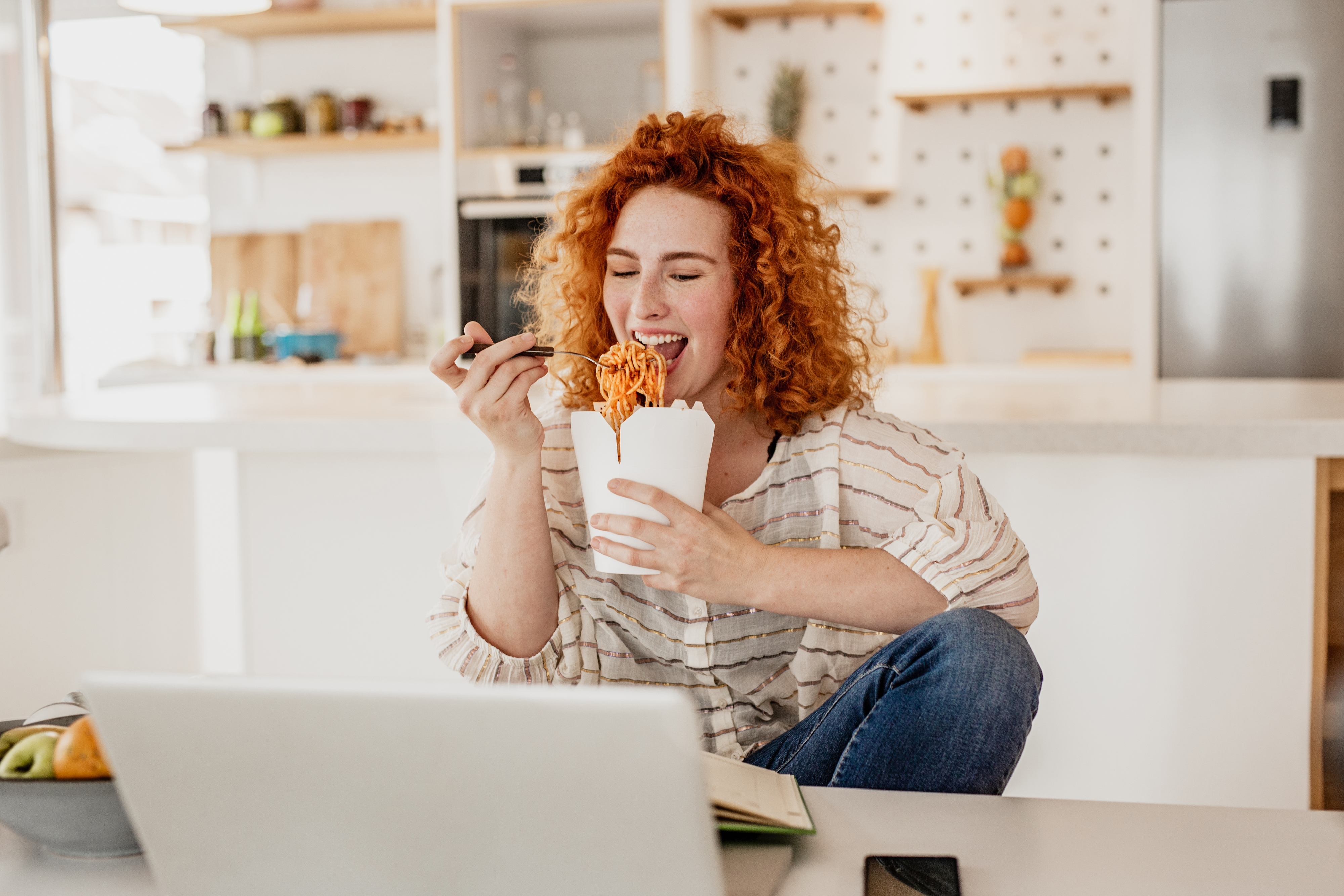 woman eating chinese takeout while smiling at her laptop and a book