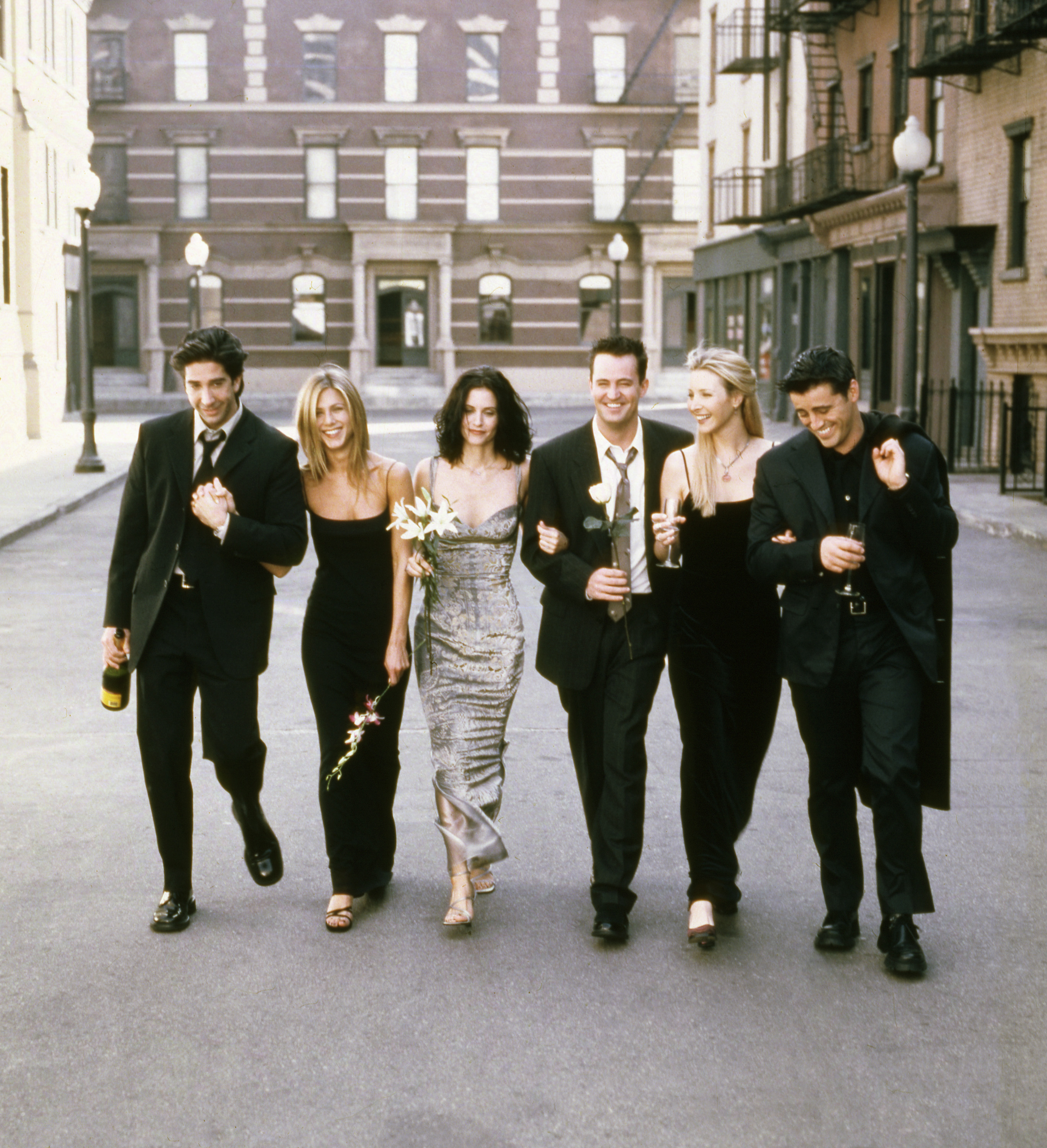 The six castmates walking down the street arm in arm, some holding champagne glasses