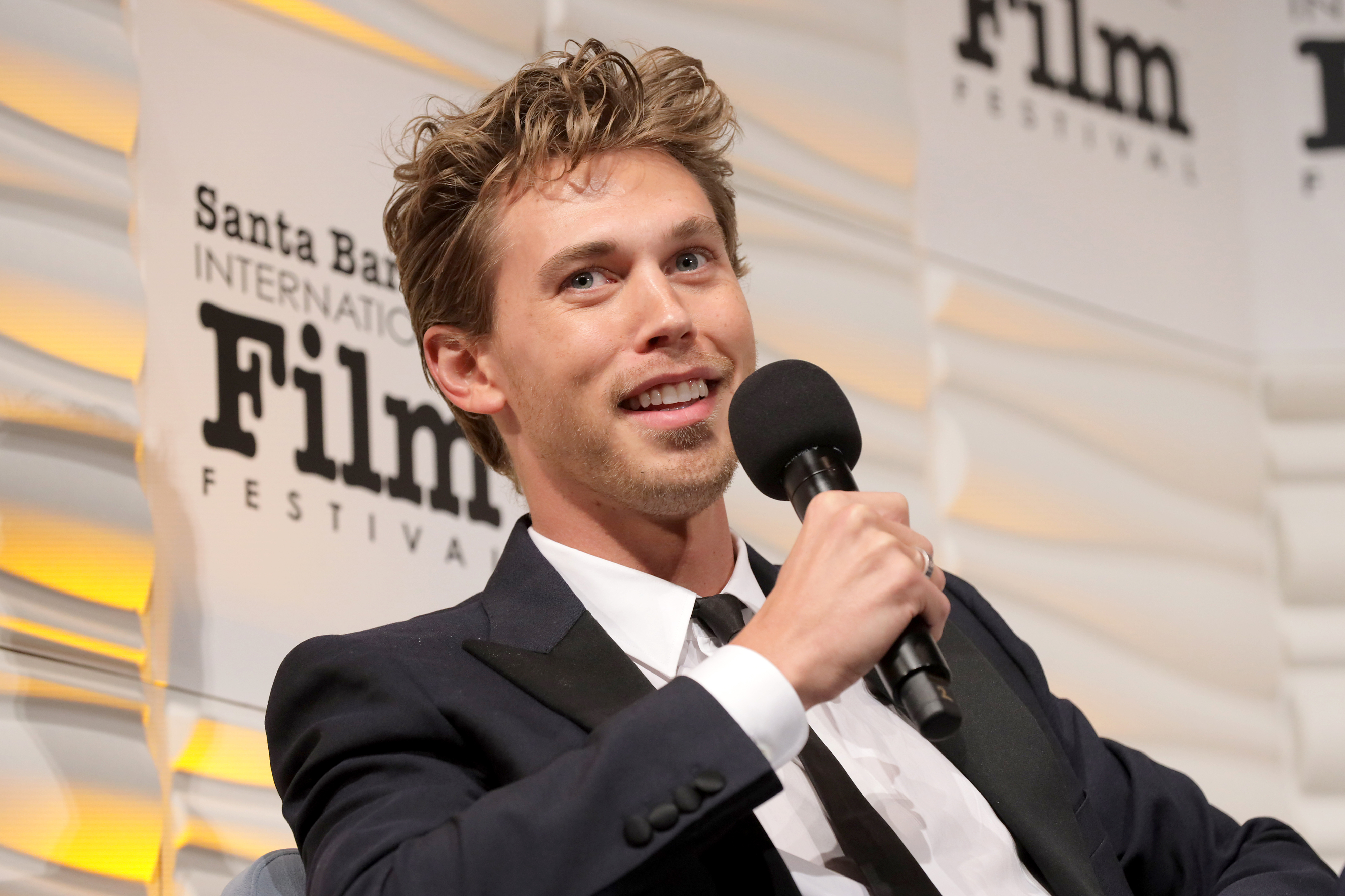 Close-up of Austin sitting onstage in a suit and tie at a media event