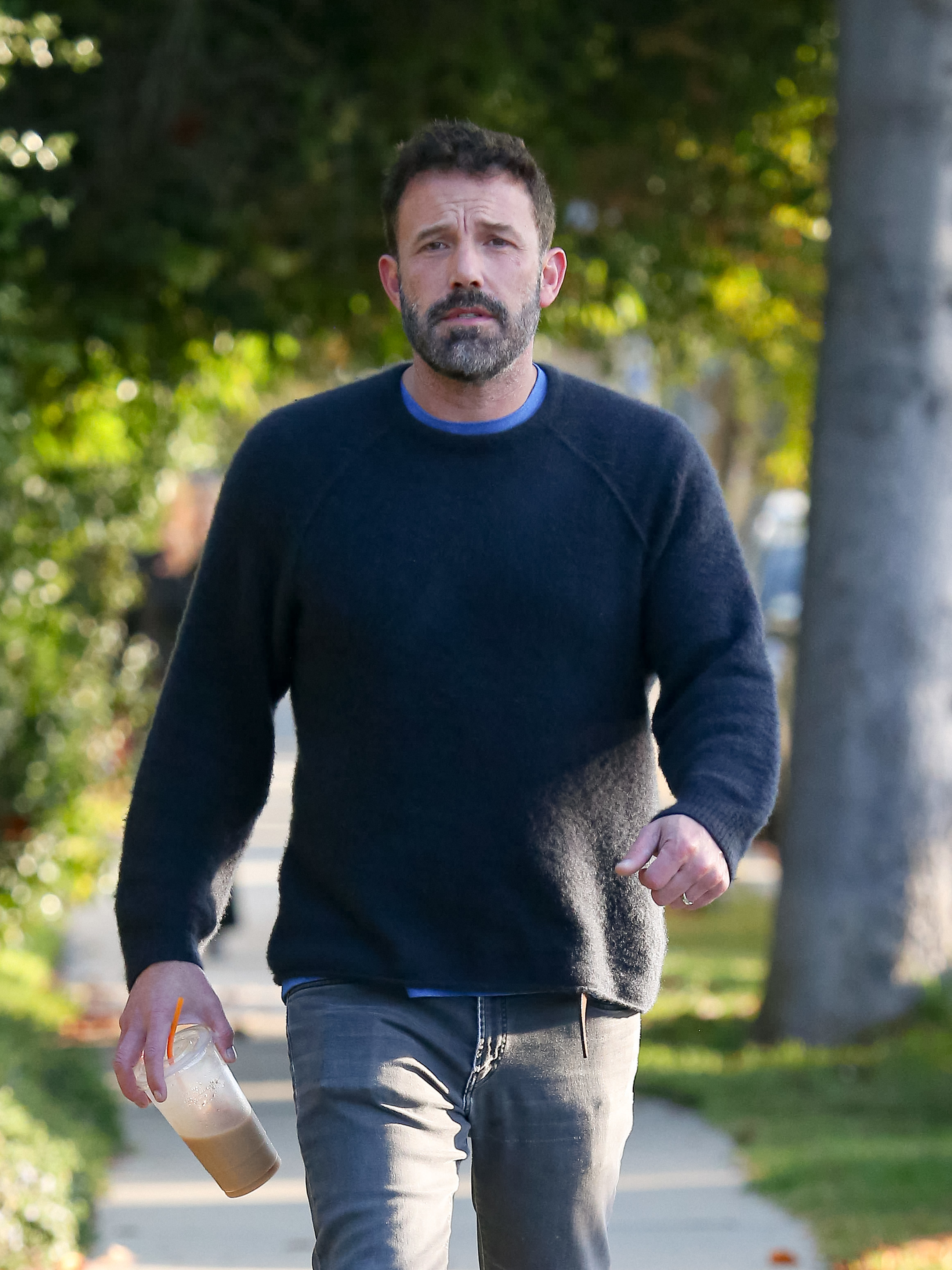 Close-up of Ben walking on a path in a sweater and jeans and wearing a takeout iced coffee