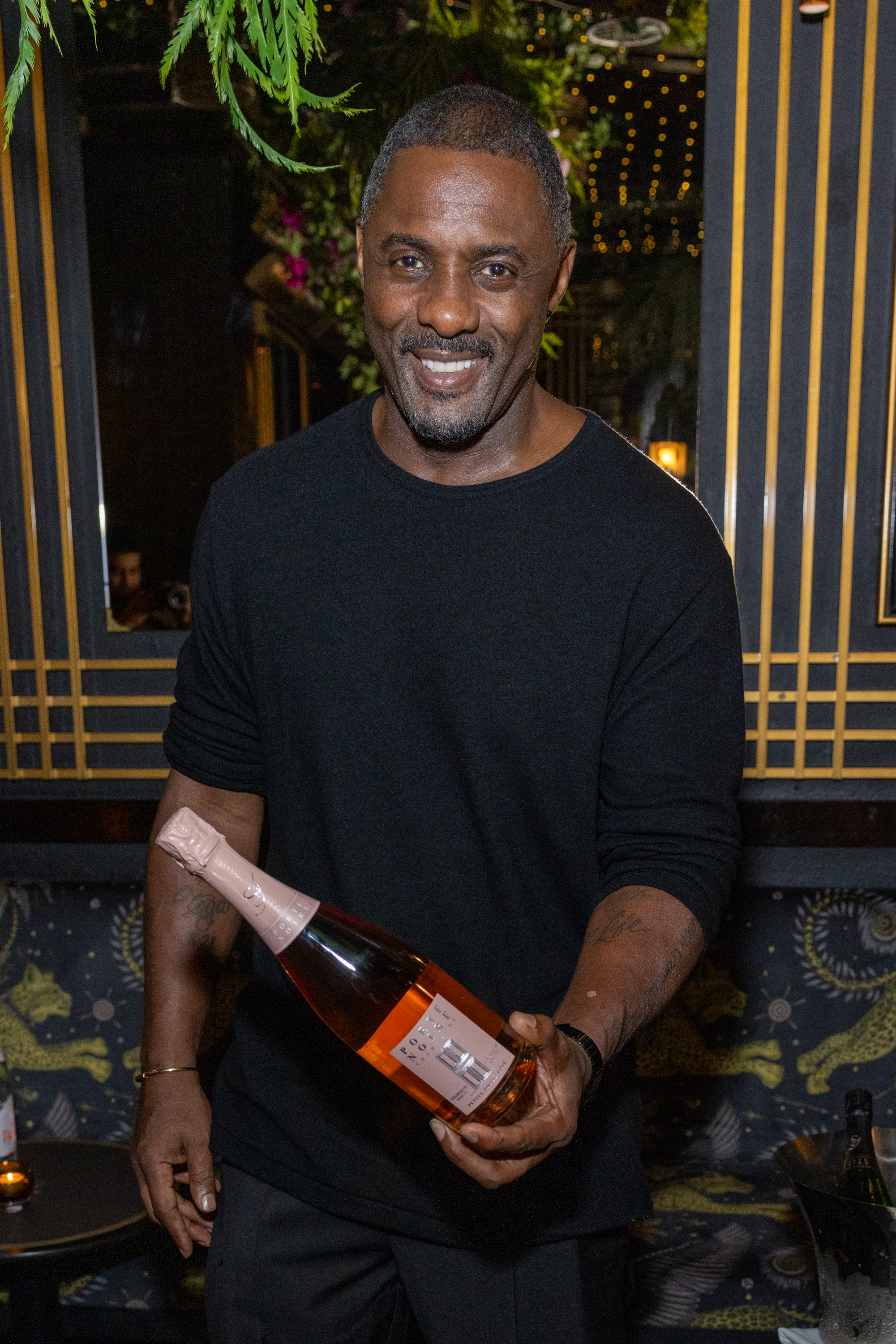 Close-up of Idris smiling in a T-shirt and holding a bottle of wine