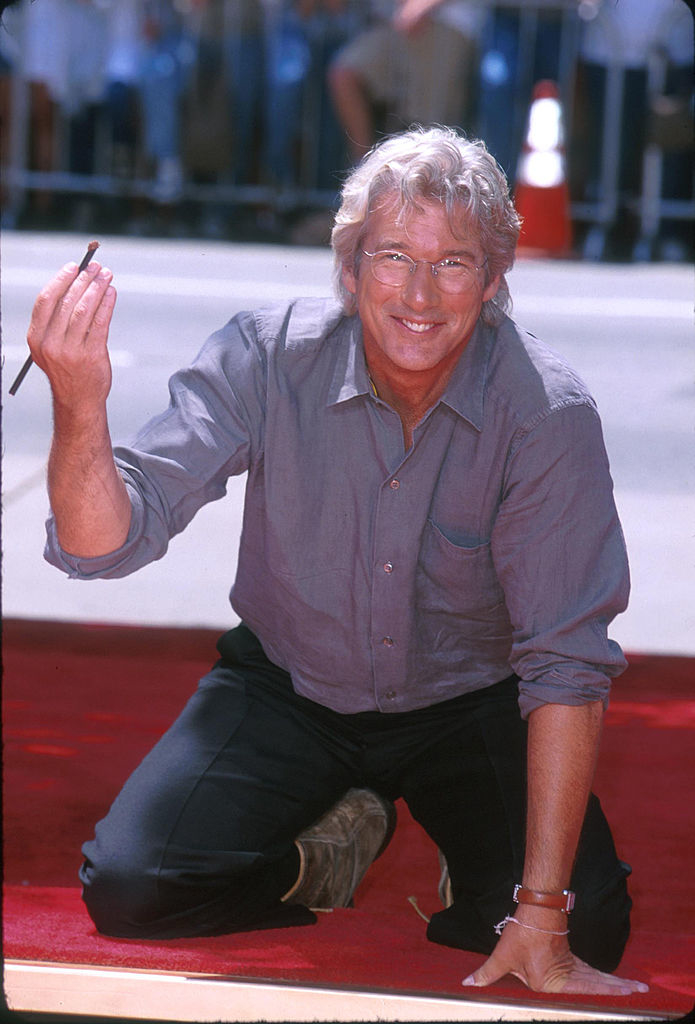 Close-up of Richard smiling and crouching on a red carpet wearing a shirt with rolled-up sleeves and pants
