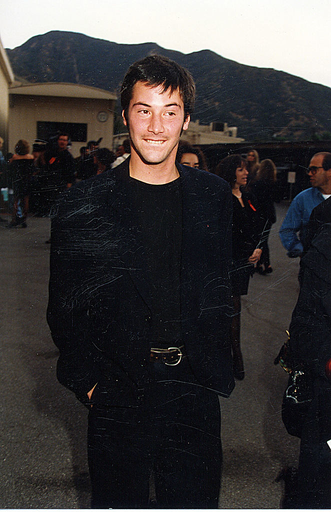 Close-up of Keanu smiling and wearing a black suit, top, and pants