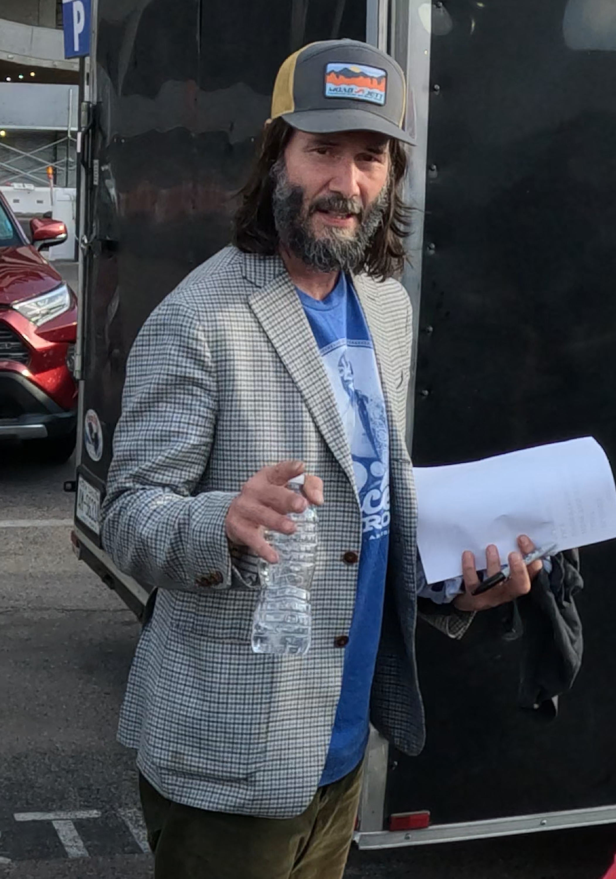 Close-up of Keanu with a mustache and heavy beard and wearing a cap, suit jacket, T-shirt, and pants