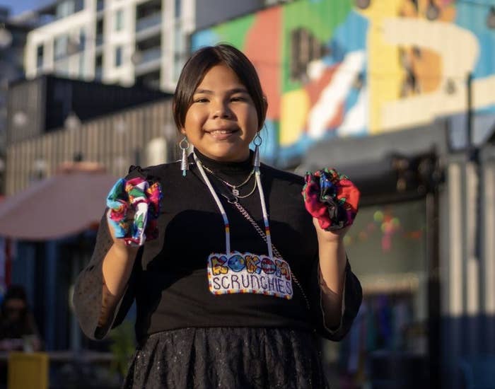 Founder of Kokom Scrunchies, Mya, holds her scrunchies  in each hand and smiles with a necklace that reads &quot;KOKUM SCRUNCHIES.&quot;