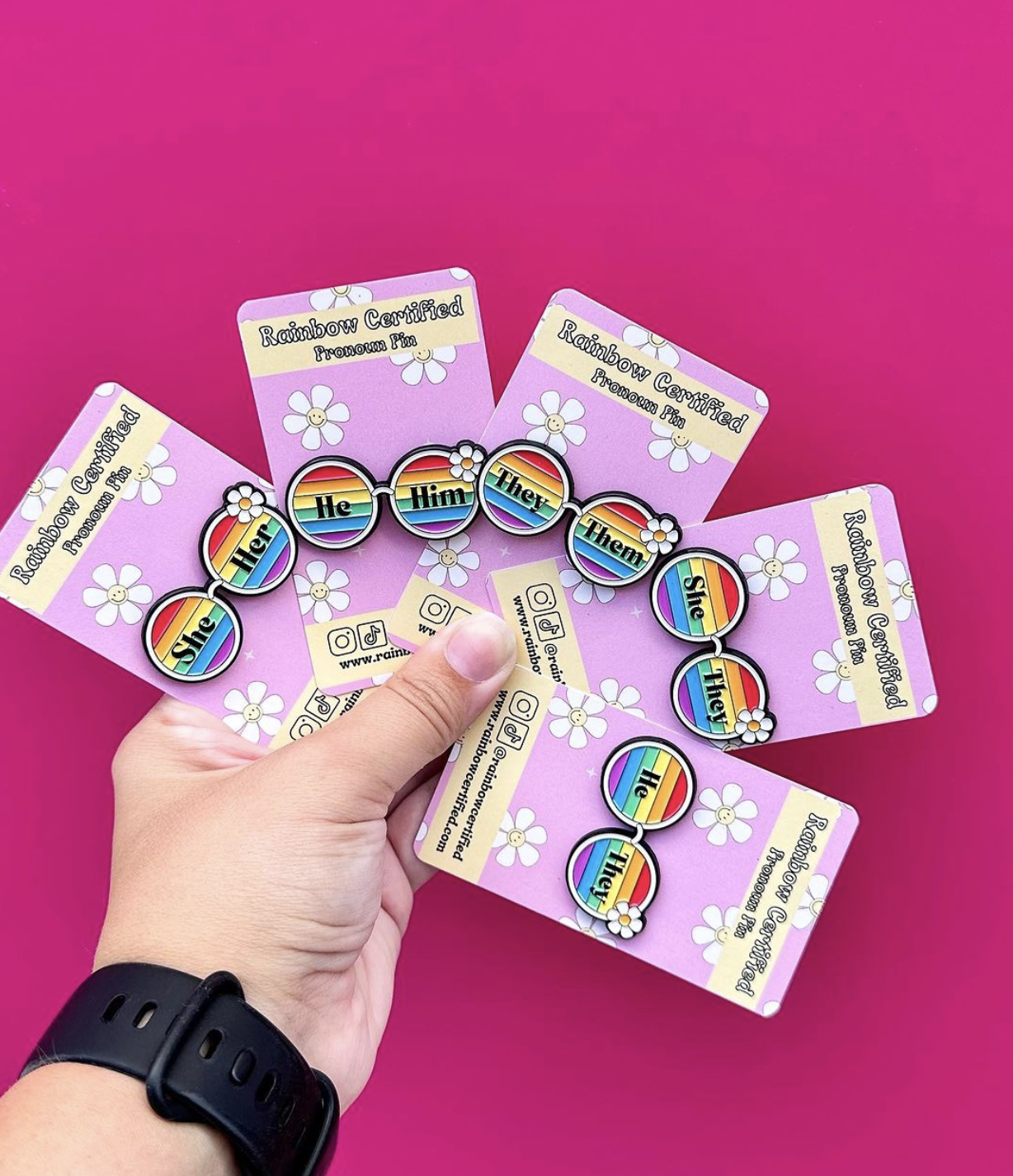 Hand holds out 4 packages of pins in a fan-like shape. Pins are circular and have different pronouns on them.