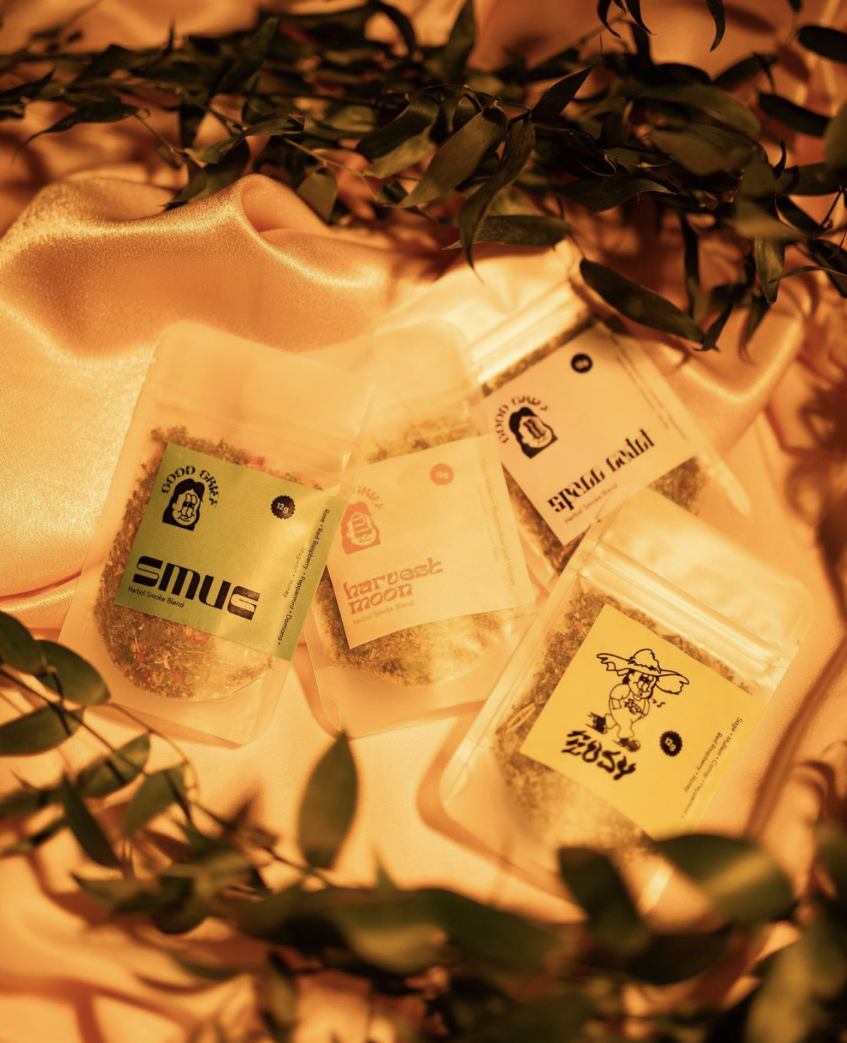 4 bags of herbal blends are posed on a silk cloth with leaves around in warm lighting.