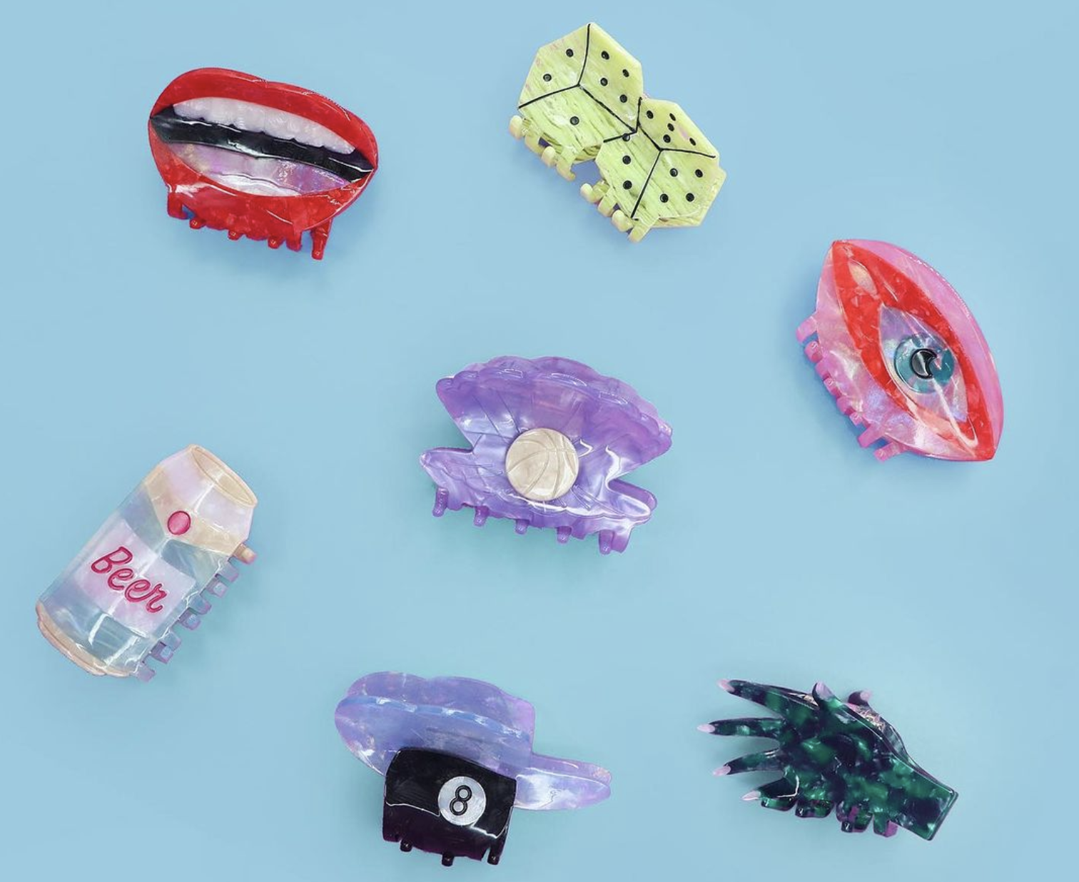 Various custom-made hair clips are laid out on a neutral background. Clips include shapes like red lips, rolled dice, 8-ball, evil eye, etc.