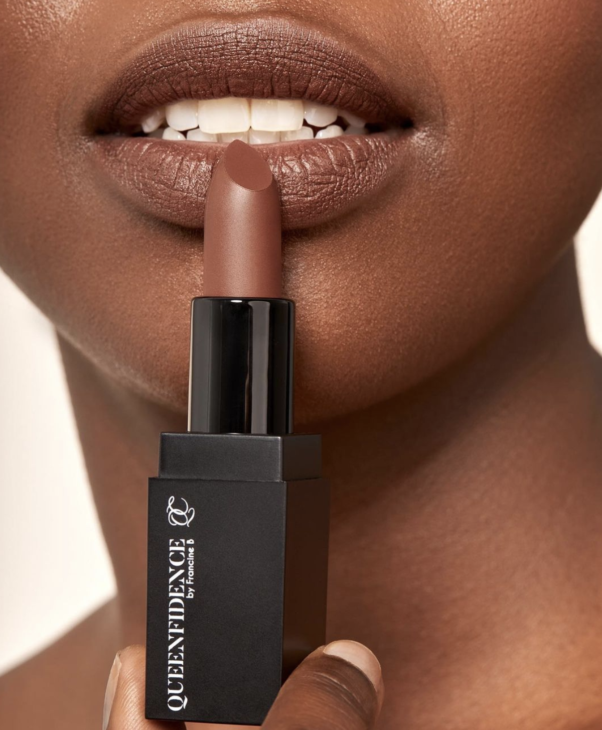 Black woman holds a brown lipstick against her lips. The lipstick container reads &quot;QUEENFIDENCE.&quot;