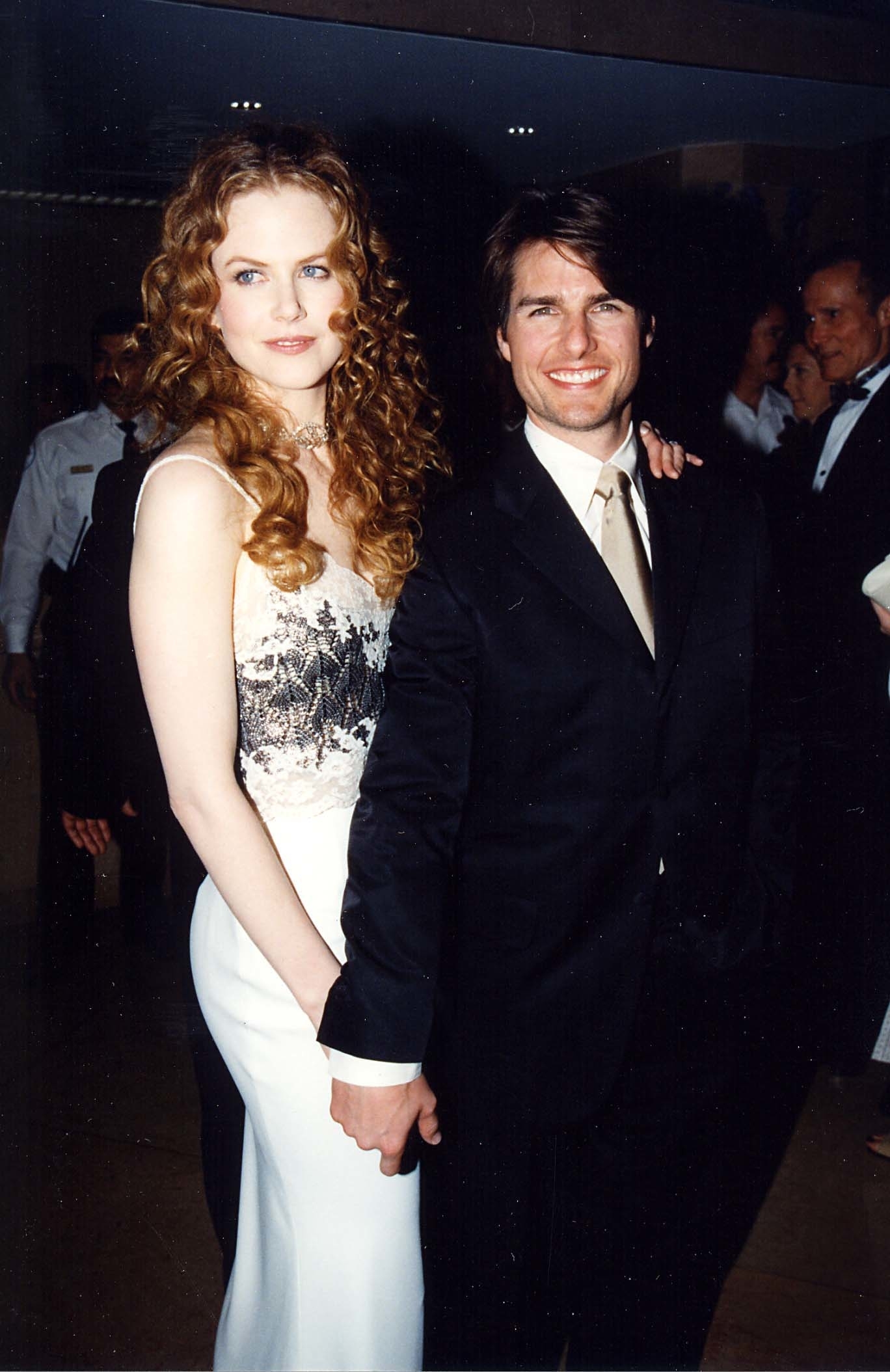 Tom in a suit and tie, smiling and holding hands with Nicole Kidman