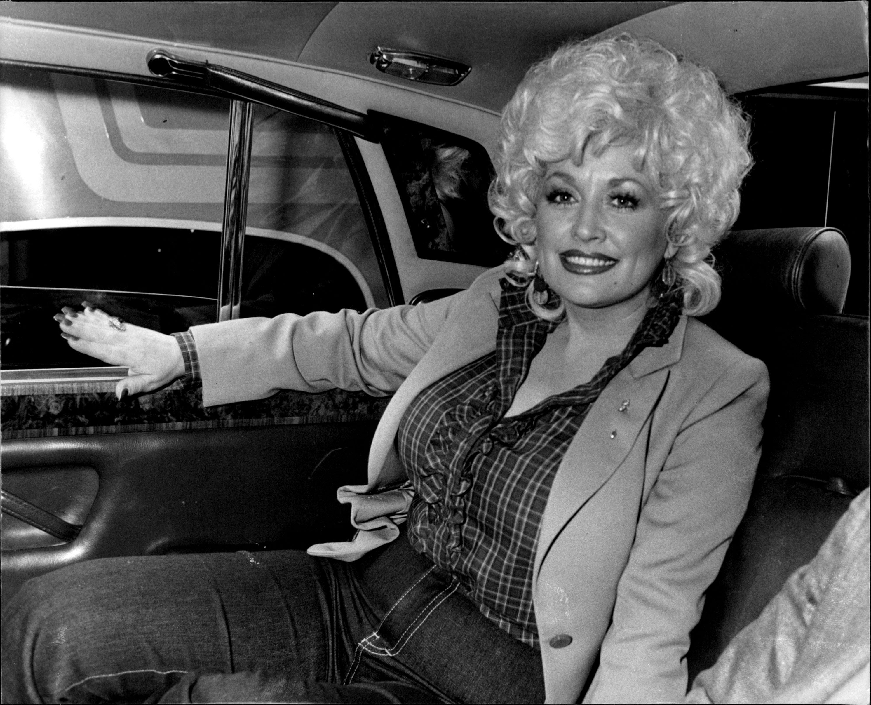 Dolly in a car, smiling and wearing jeans, a plaid blouse, and a jacket