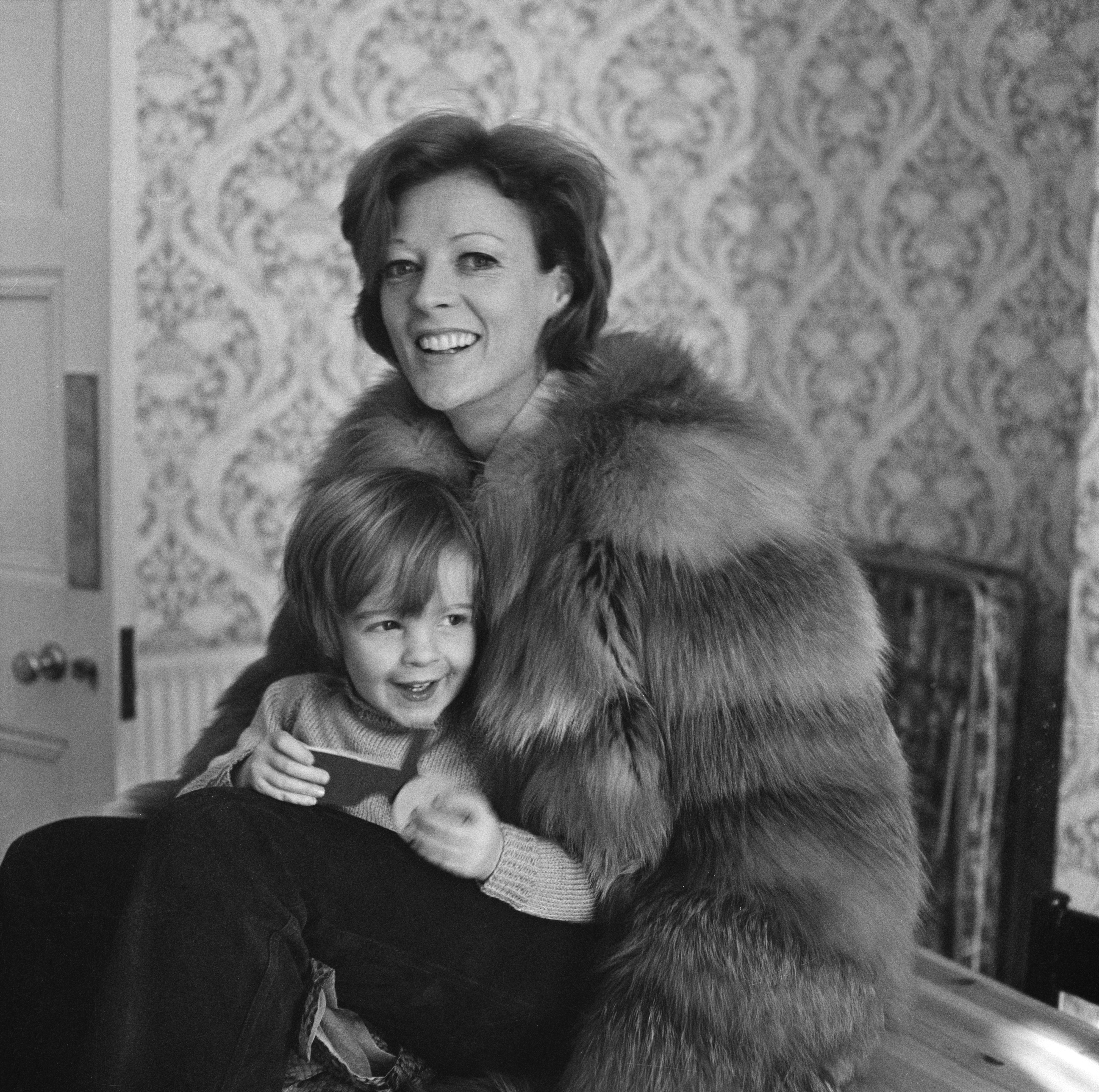 Maggie smiling, sitting with a young child, and wearing a fur coat and pants