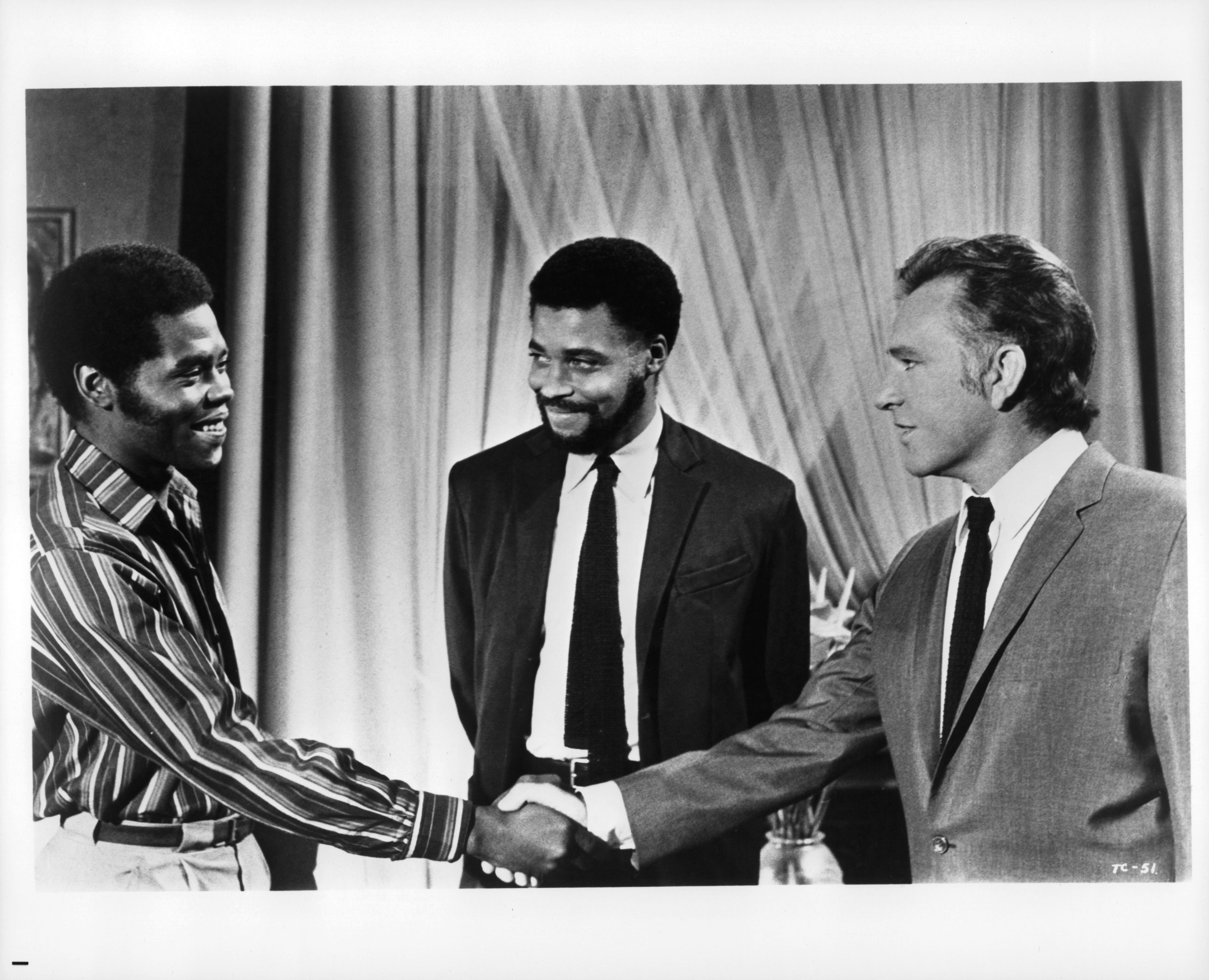 James standing between Richard Burton and Georg Stanford Brown, who are shaking hands