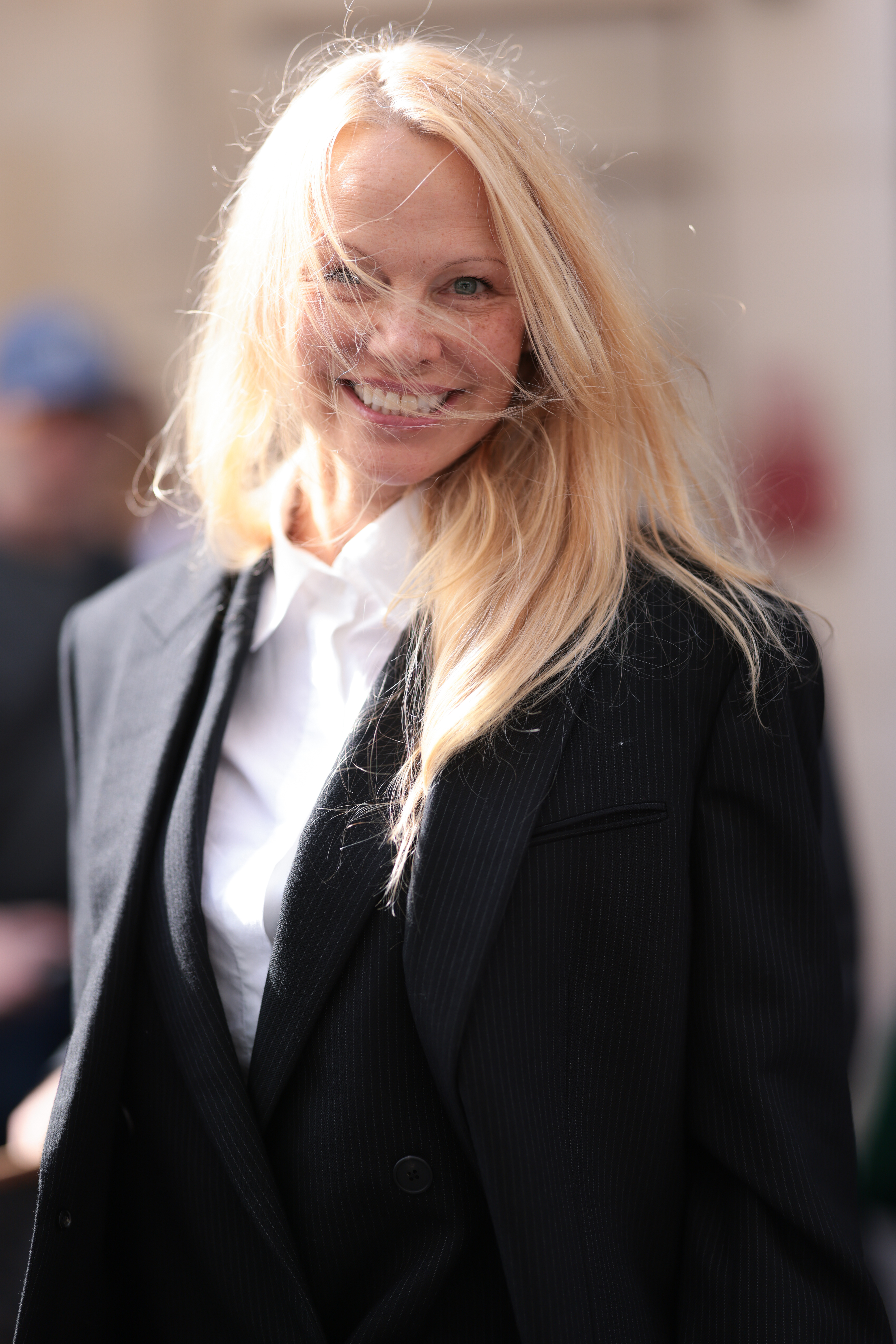 Closeup of Pamela Anderson smiling widely as she walks wearing a blouse and a blazer