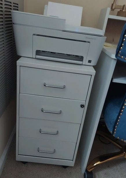 A grey printer sits on top of a filing cabinet with four drawers in an office setting. There&#x27;s no visible text or people in the image