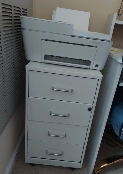 A grey printer sits on top of a filing cabinet with four drawers in an office setting. There's no visible text or people in the image