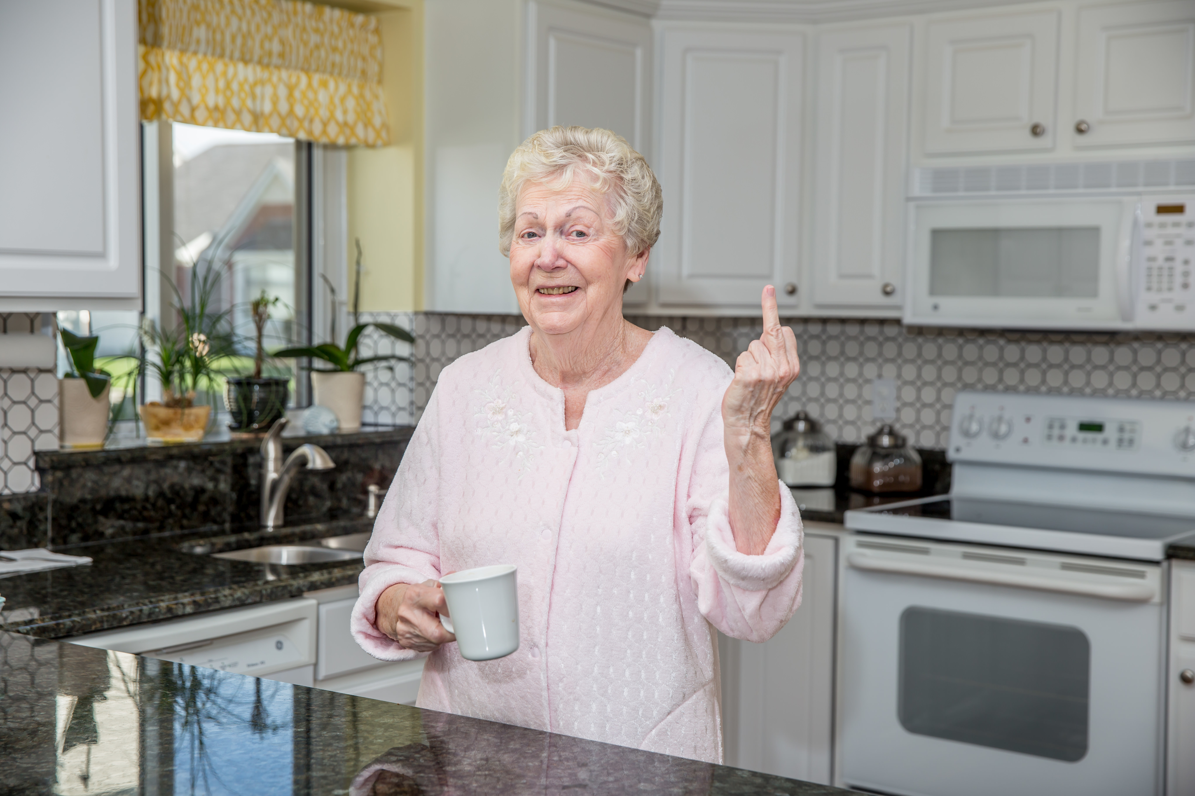 An older woman in a kitchen holding a mug, smiling, and raising her middle finger