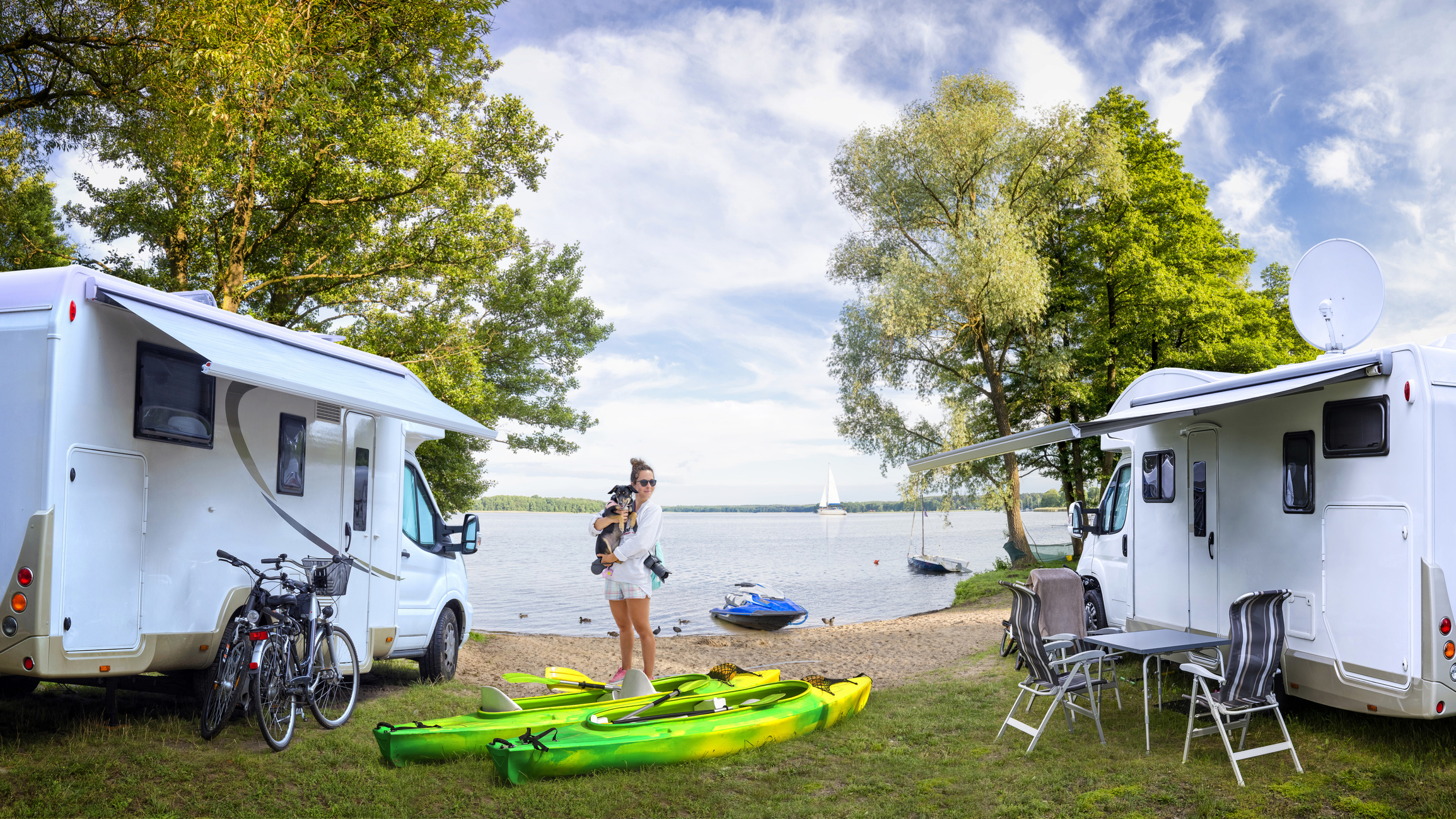 A woman holding her dog outside her camper in front of a lake with kayaks and bikes