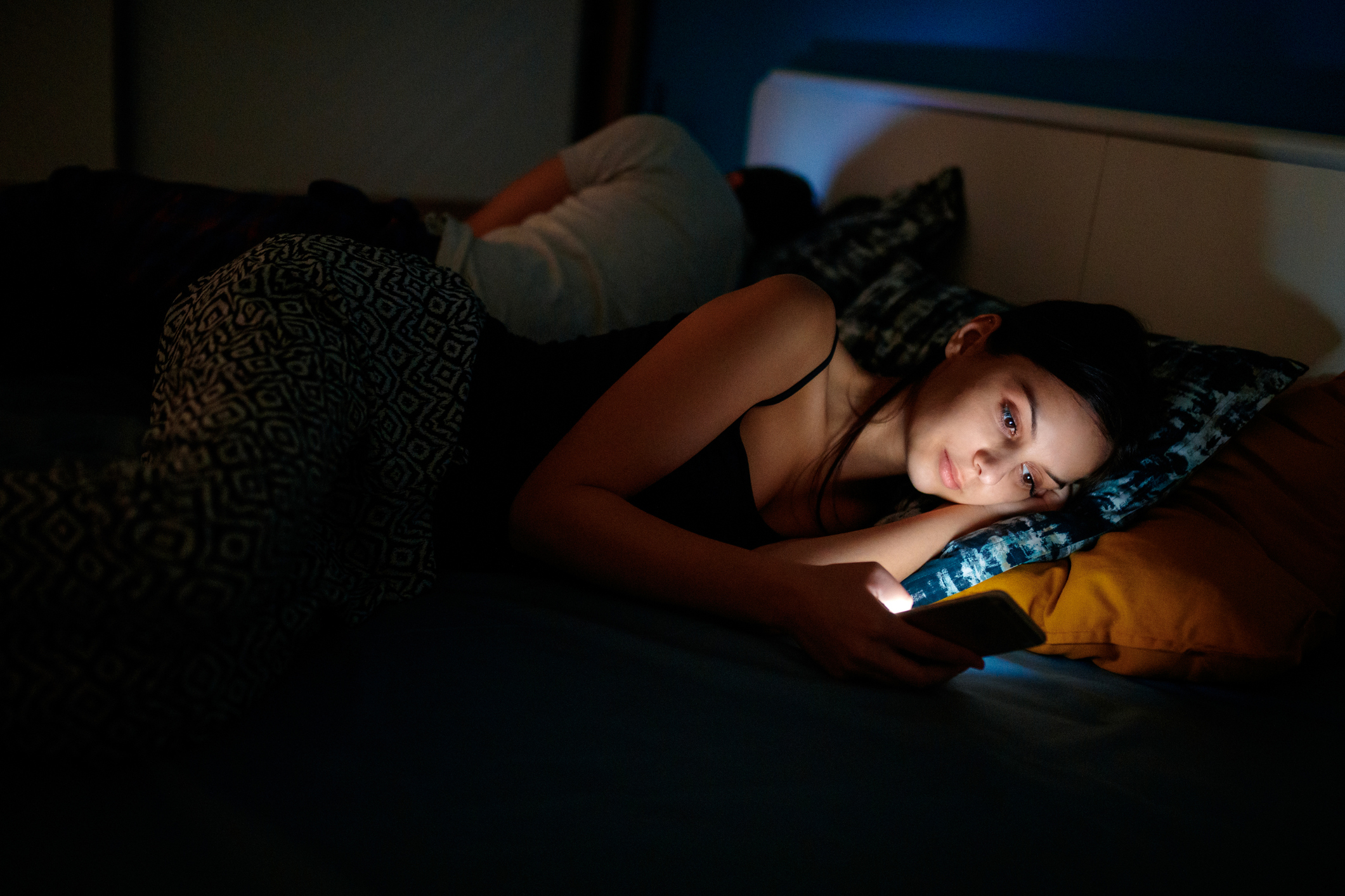 Young woman sending text message as her boyfriend sleeps in the bed, as she is turned away from him