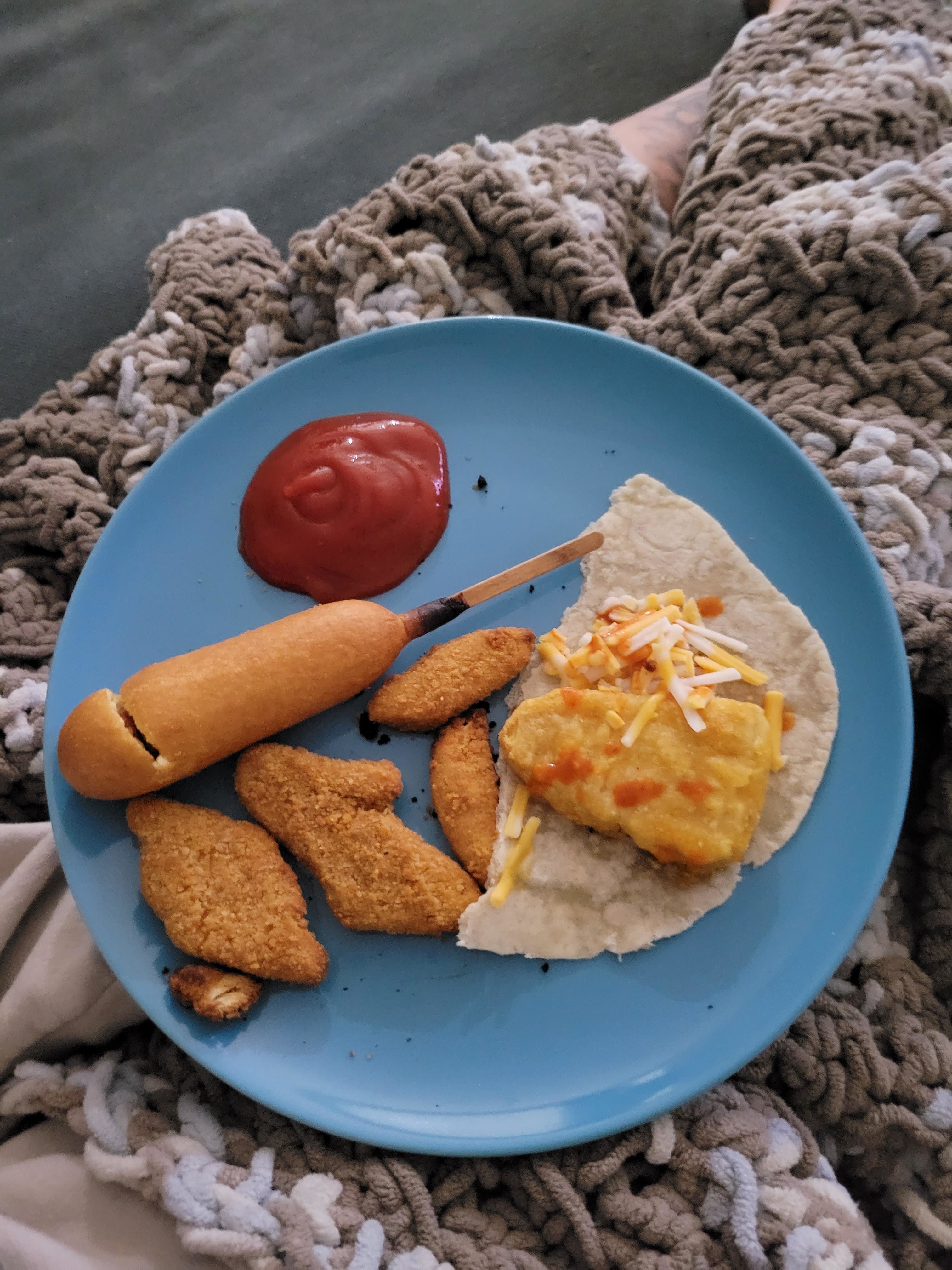 a plate with half a tortilla, a frozen fish piece, four chicken tenders, a broken corn dog, and some ketchup