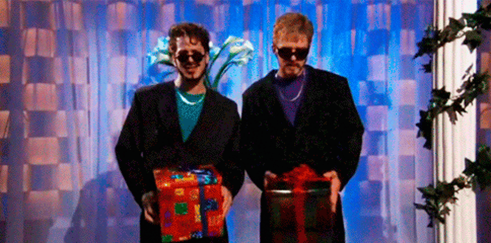 Two men in suits holding gift boxes near their waists on a stage.