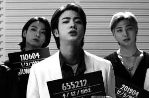 Three of the BTS boys stand in a lineup, getting mugshots taken, in a music video scene.