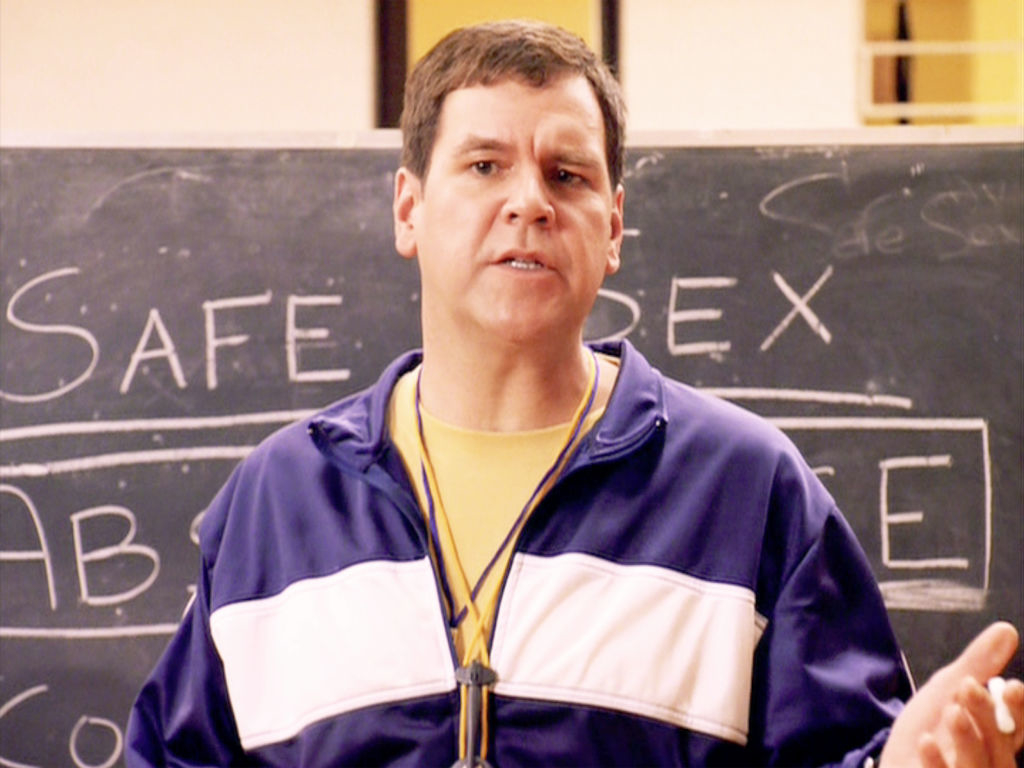 closeup of him in a track jacket in front of a chalk board that says, safe sex