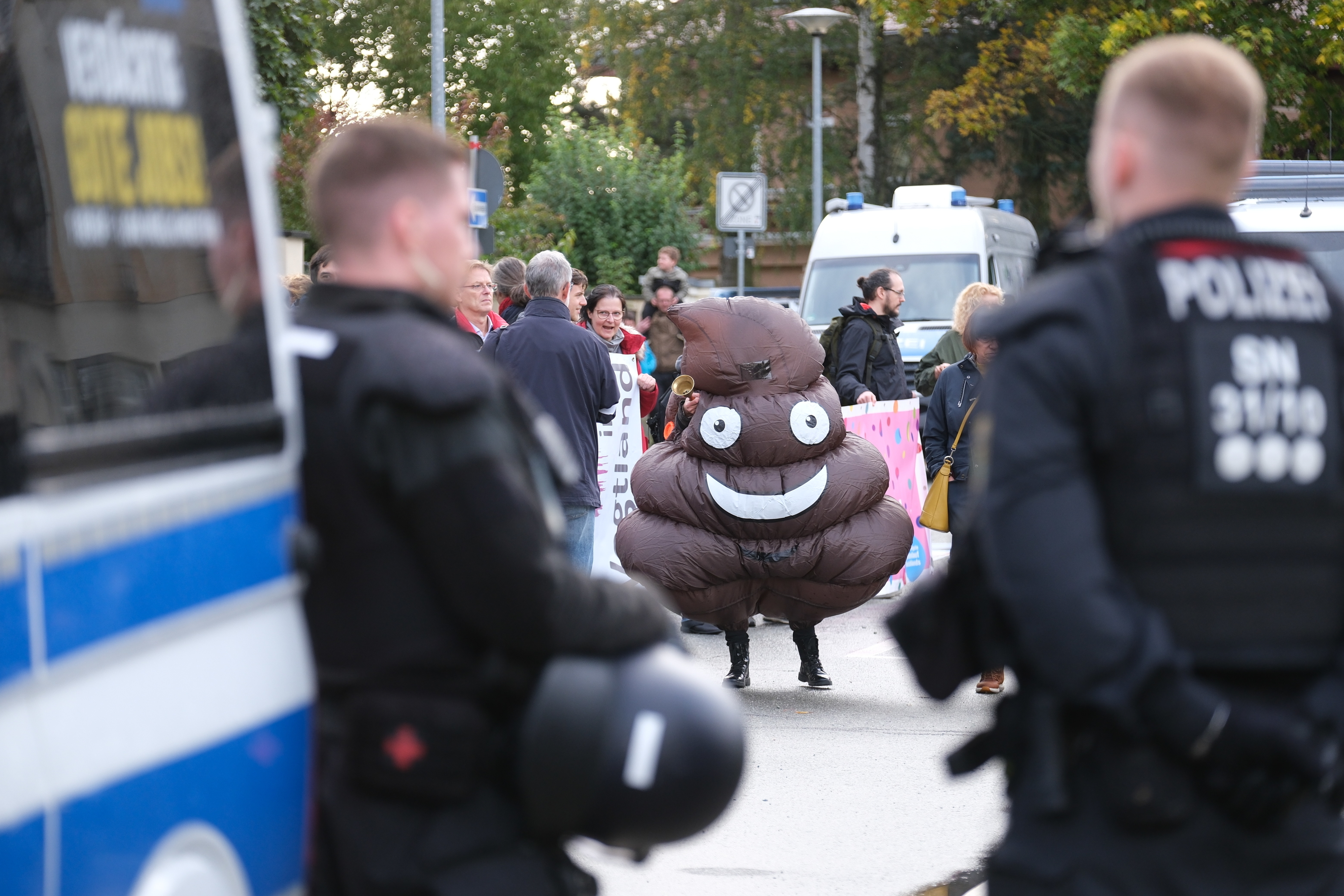 a person dressed as the poop emoji (a swirly piece of poop with eyes) stands in the background, between two blurry police officers in the foreground