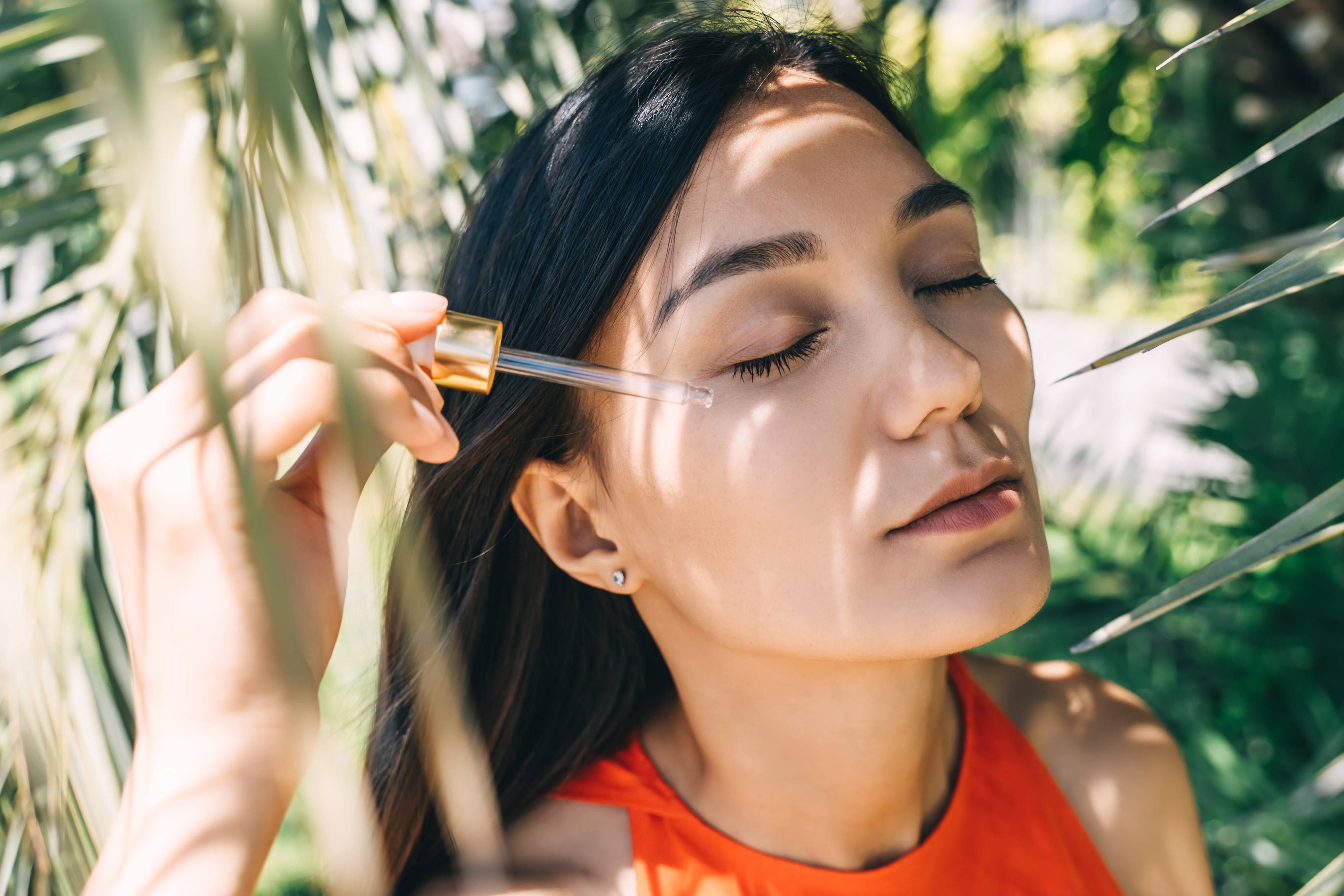 A beautiful young woman applies serum to her face with a pipette. Close-up portrait between palm leaves.