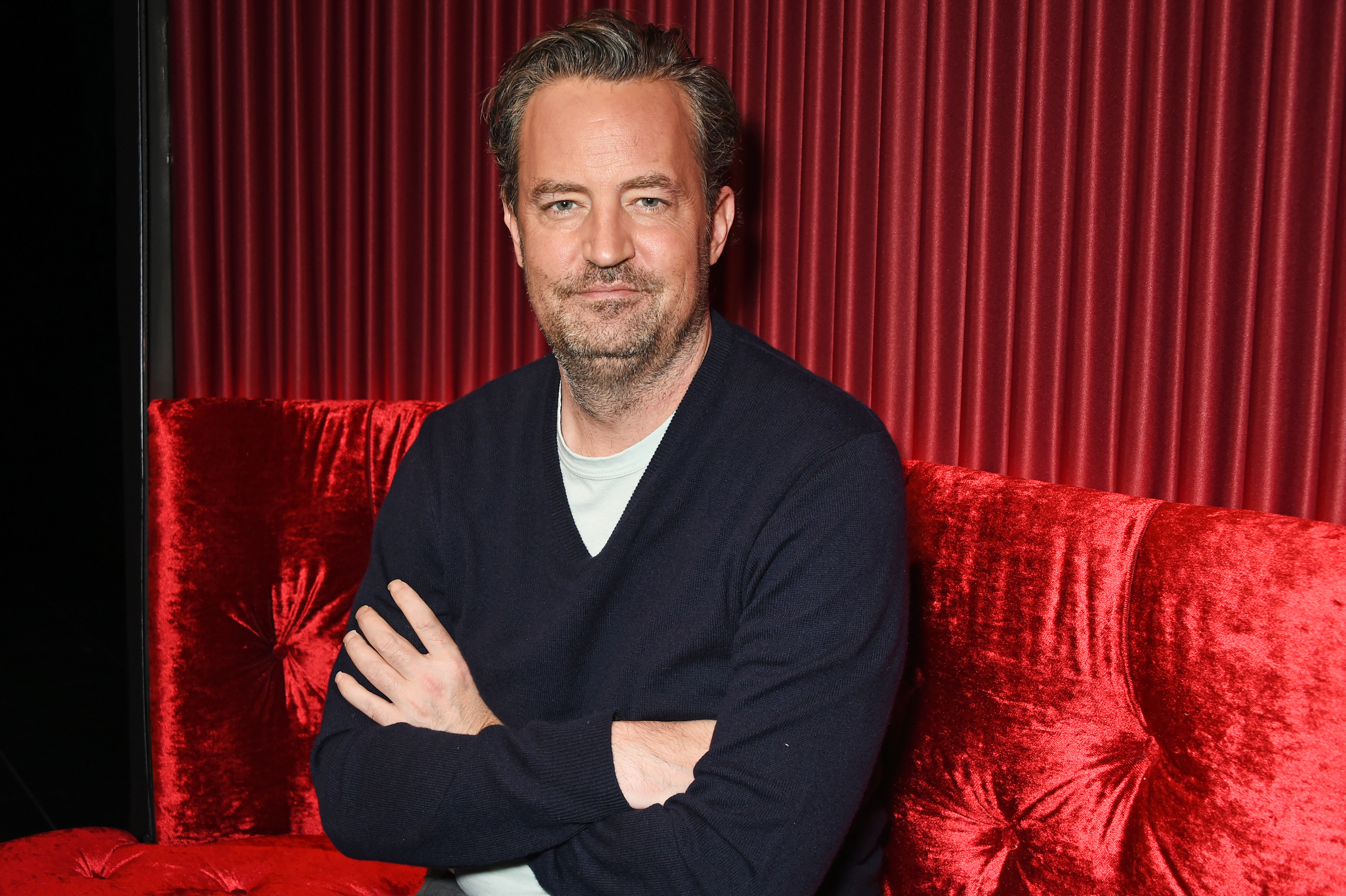 Matthew Perry sitting with his arms crossed on a velvet couch