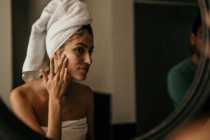 A young woman applies moisturizer to her face, her reflection in the mirror capturing her dedication to self-care.
