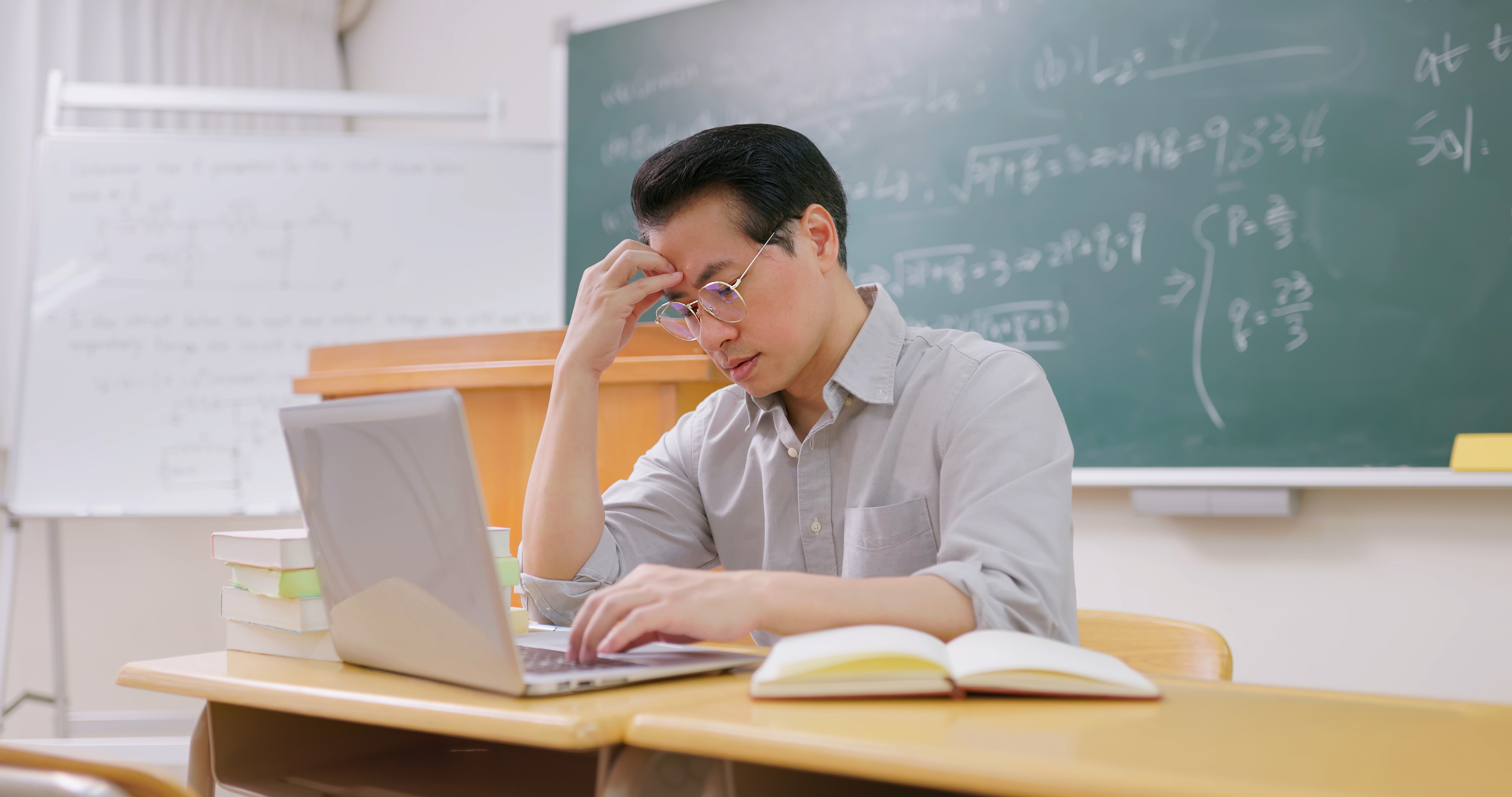 a teacher looking at a laptop holds his hand to his forehead in frustration