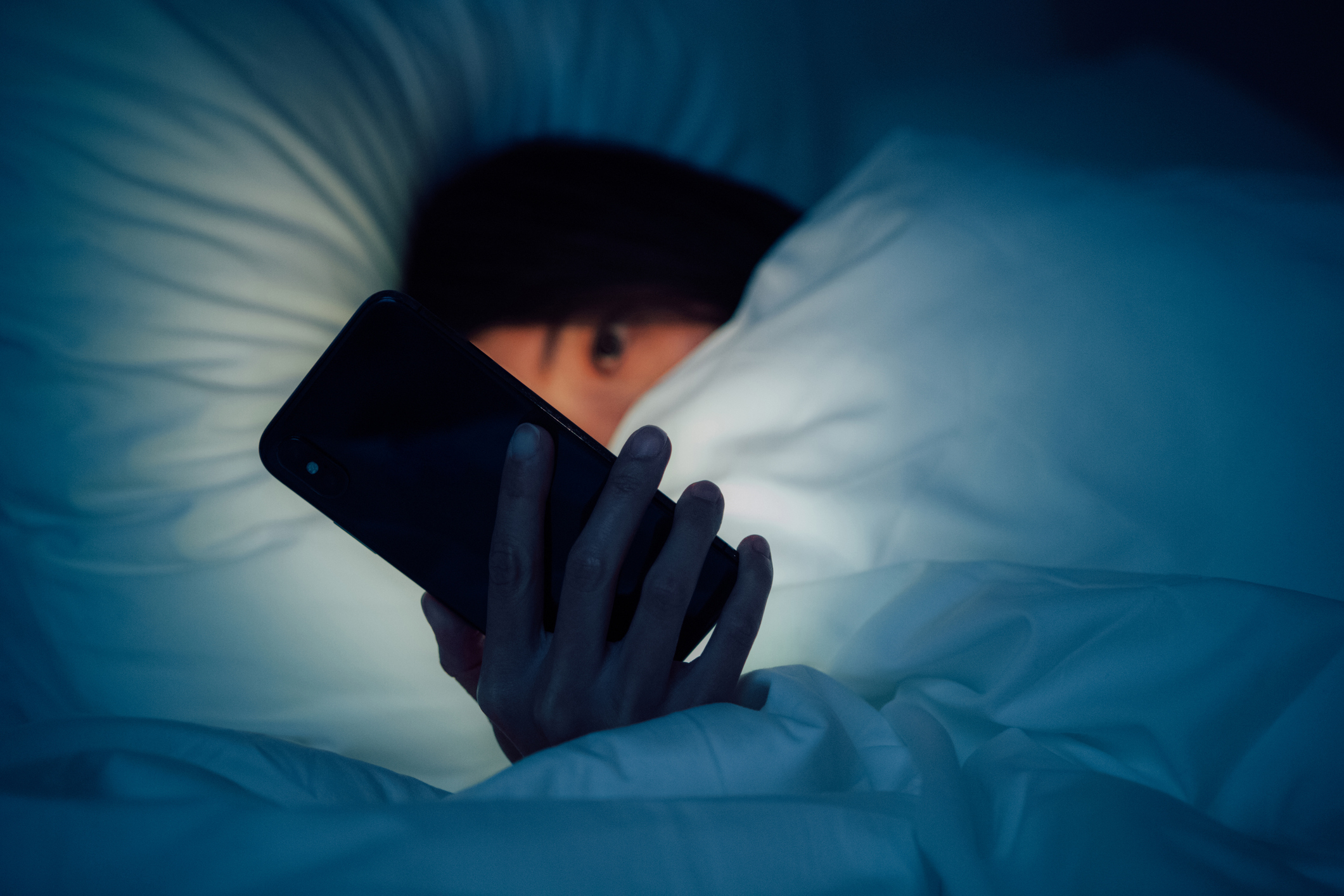 A woman peering out from under her covers in bed to look at her phone screen, which is illuminating her face in a swath of light from the screen