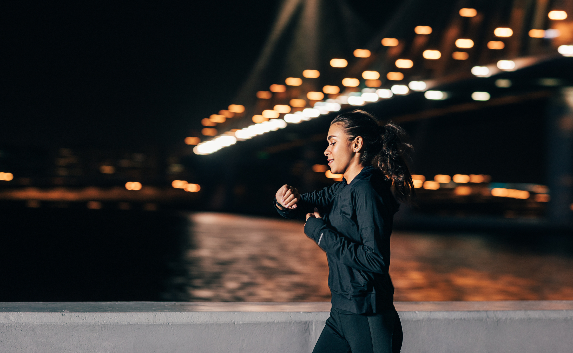 A young woman jogging during night, with city lights in the background, as she checks her exercise watch to see her pace
