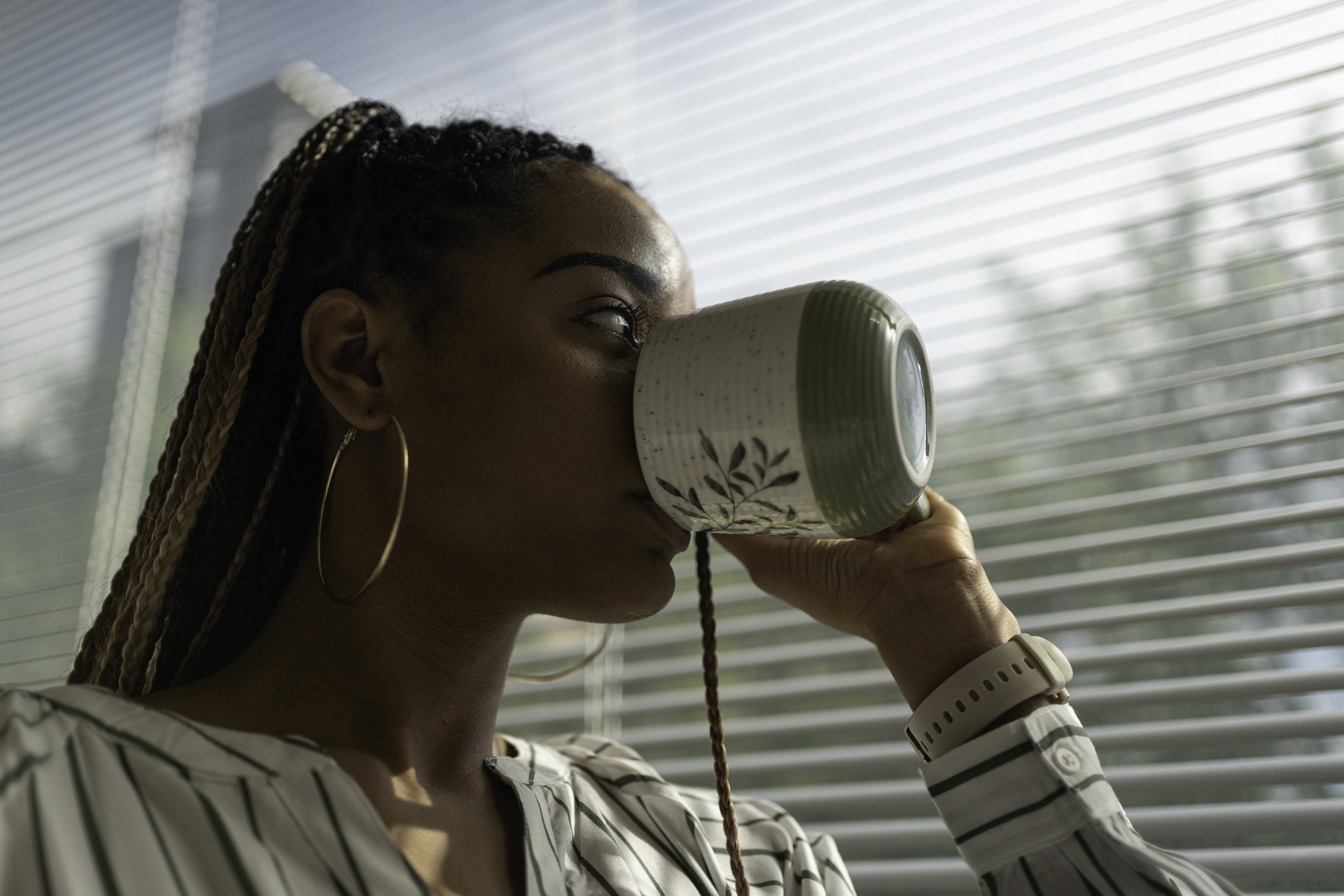 A young Black woman drinking out of a coffee cup while looking out a window