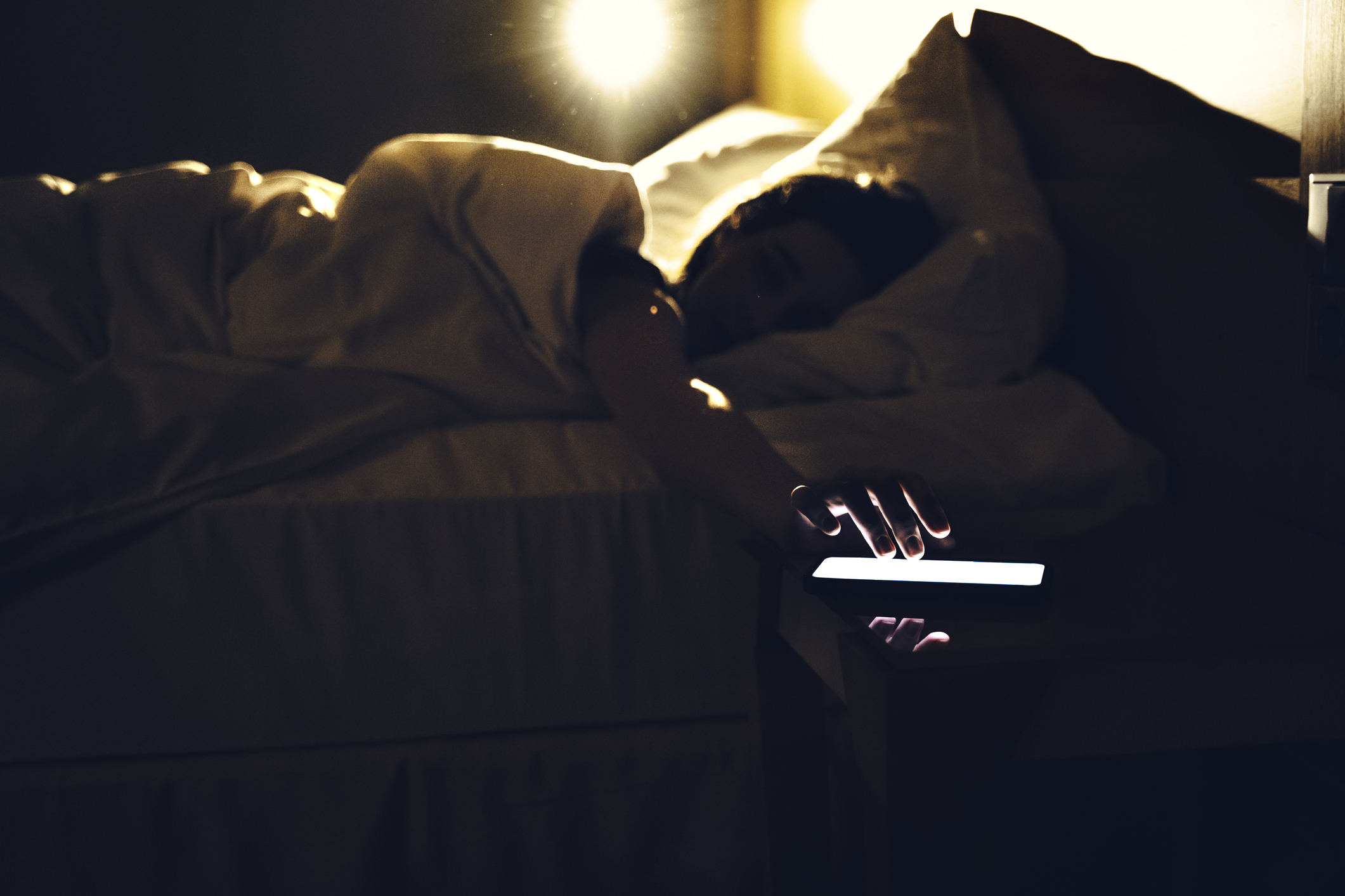 A woman reaching out from under the covers in bed to turn off the phone alarm on the side table next to the bed