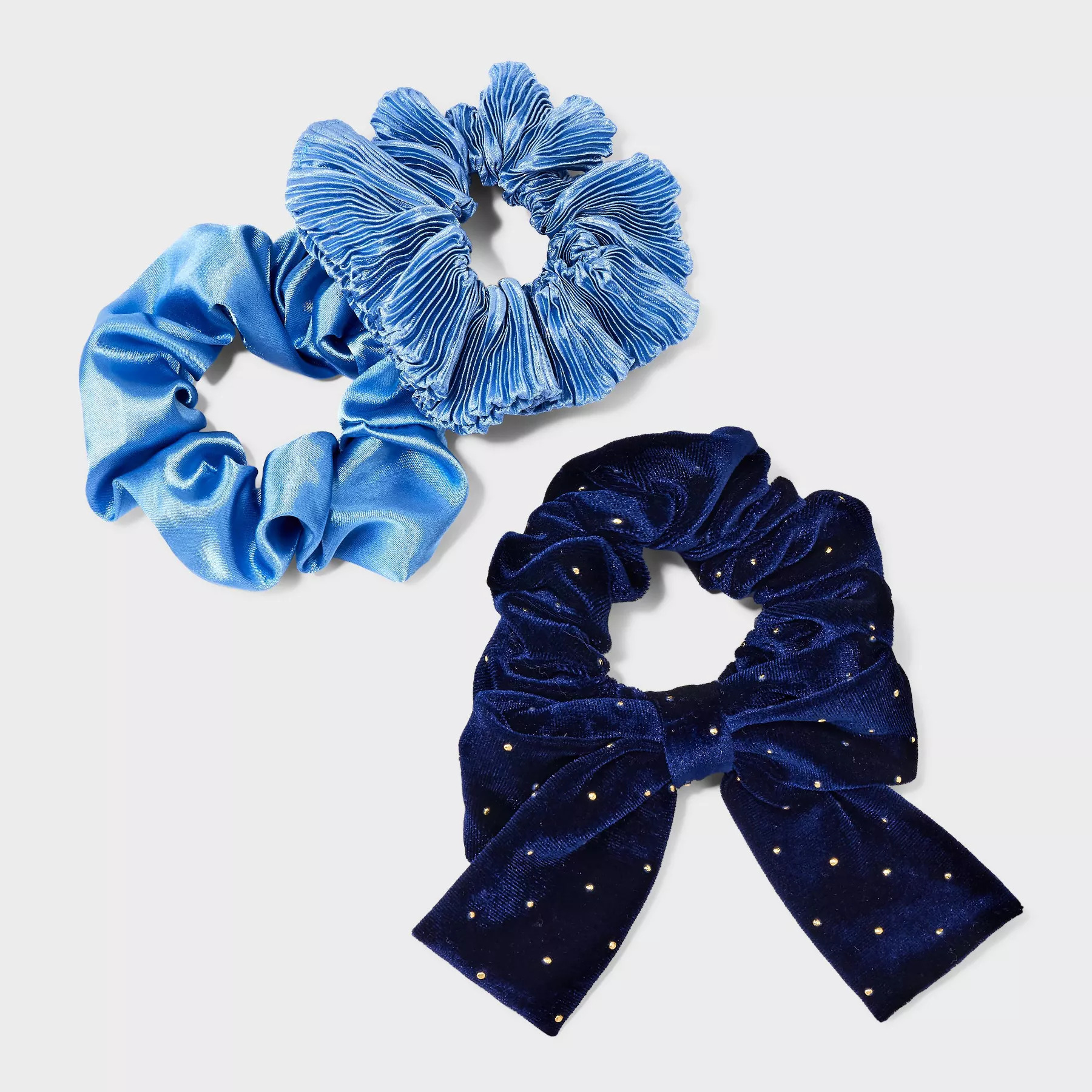The three scrunchies, one featuring a velvety finish and bow, one with a folded textured design and one with a satin finish in different shades of blue