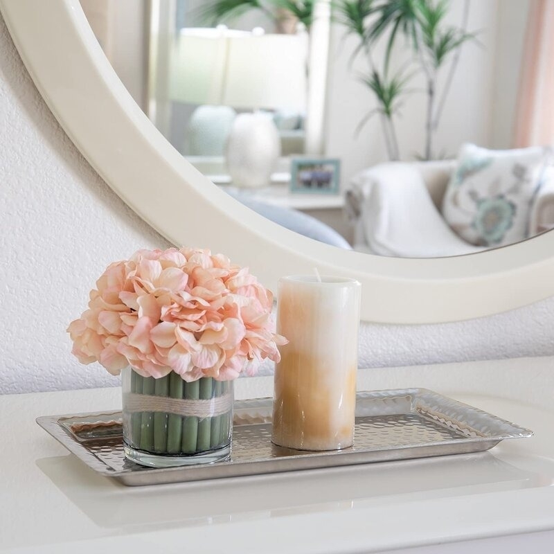 A mirrored tray displays a pink flower bouquet in a vase and a tall candle, set on a white surface in a cozy living room setting