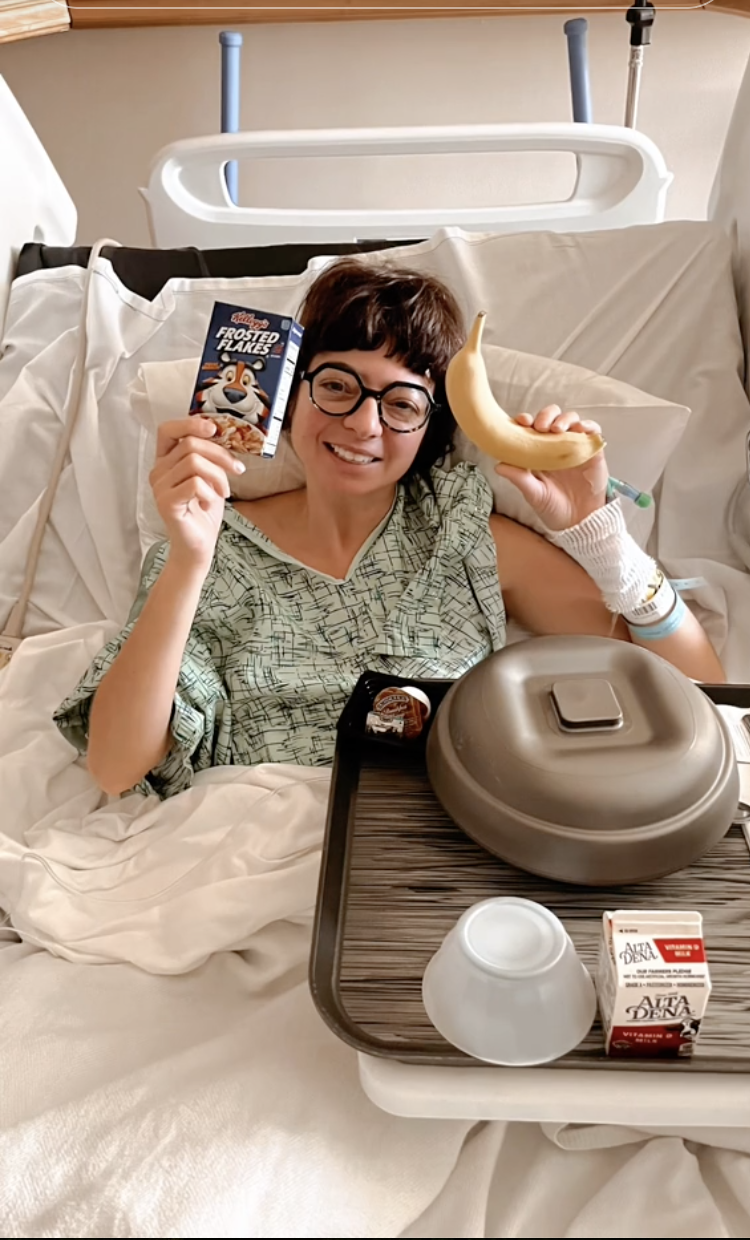 Kate Micucci in the hospital holding up a small box of cereal and a banana