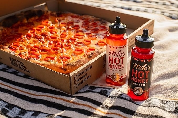a bottle of hot honey and a bottle of extra hot honey beside an open box of pizza