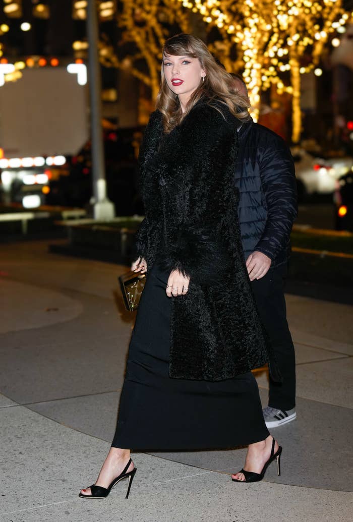 Taylor Swift walks into a building wearing a long, black fur coat.