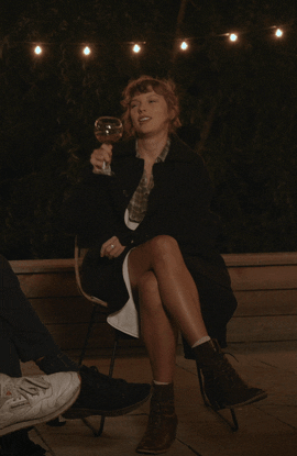 Taylor Swift sits with legs crossed and cheers' a glass of white wine.