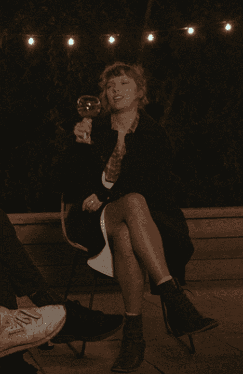 Taylor Swift sits with legs crossed and cheers&#x27; a glass of white wine.