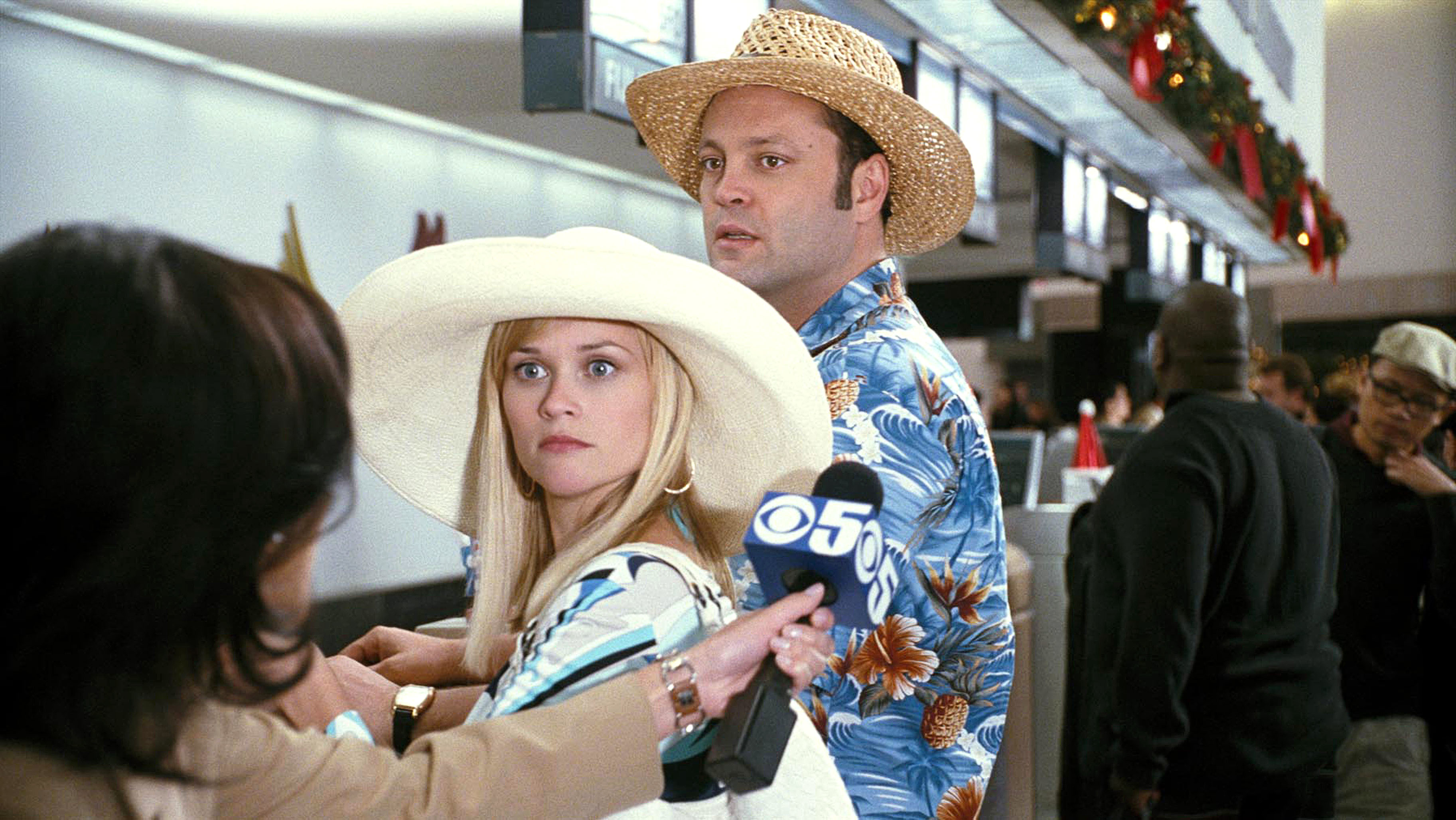 reese witherspoon and vince vaughn wearing sun hats and hawaiian shirts, being interviewed by the news at the airport