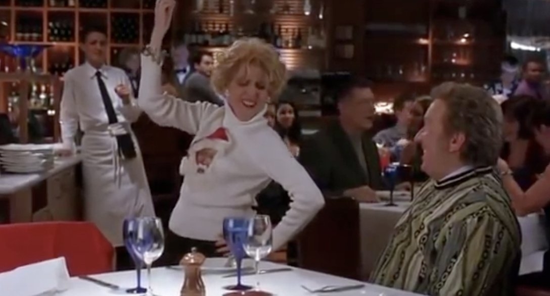 molly shannon in a christmas sweater dancing in a restaurant near her table