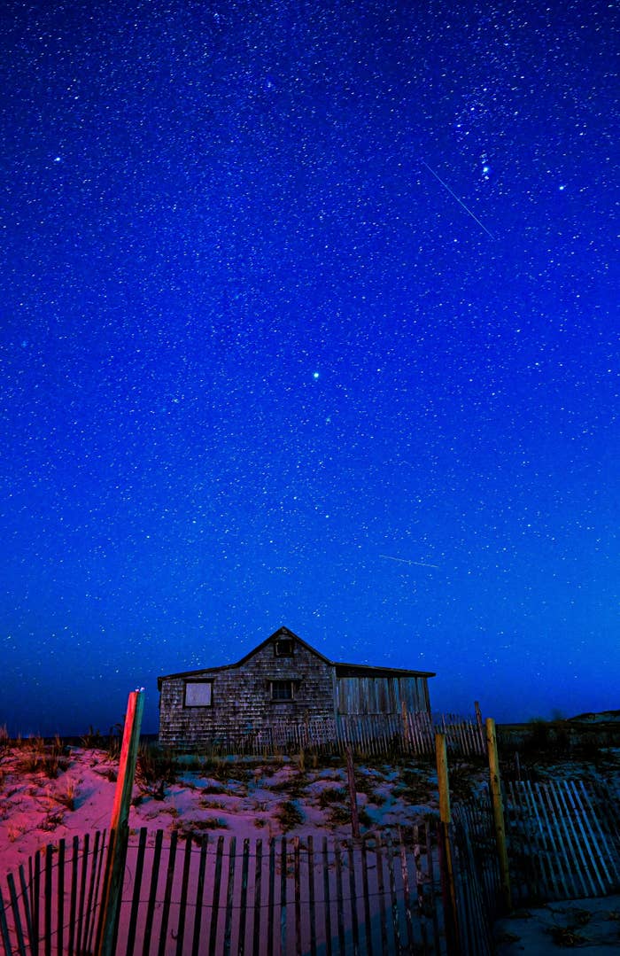View of the starry night sky above a desolate building surrounded by patches of snowy grass and a fence