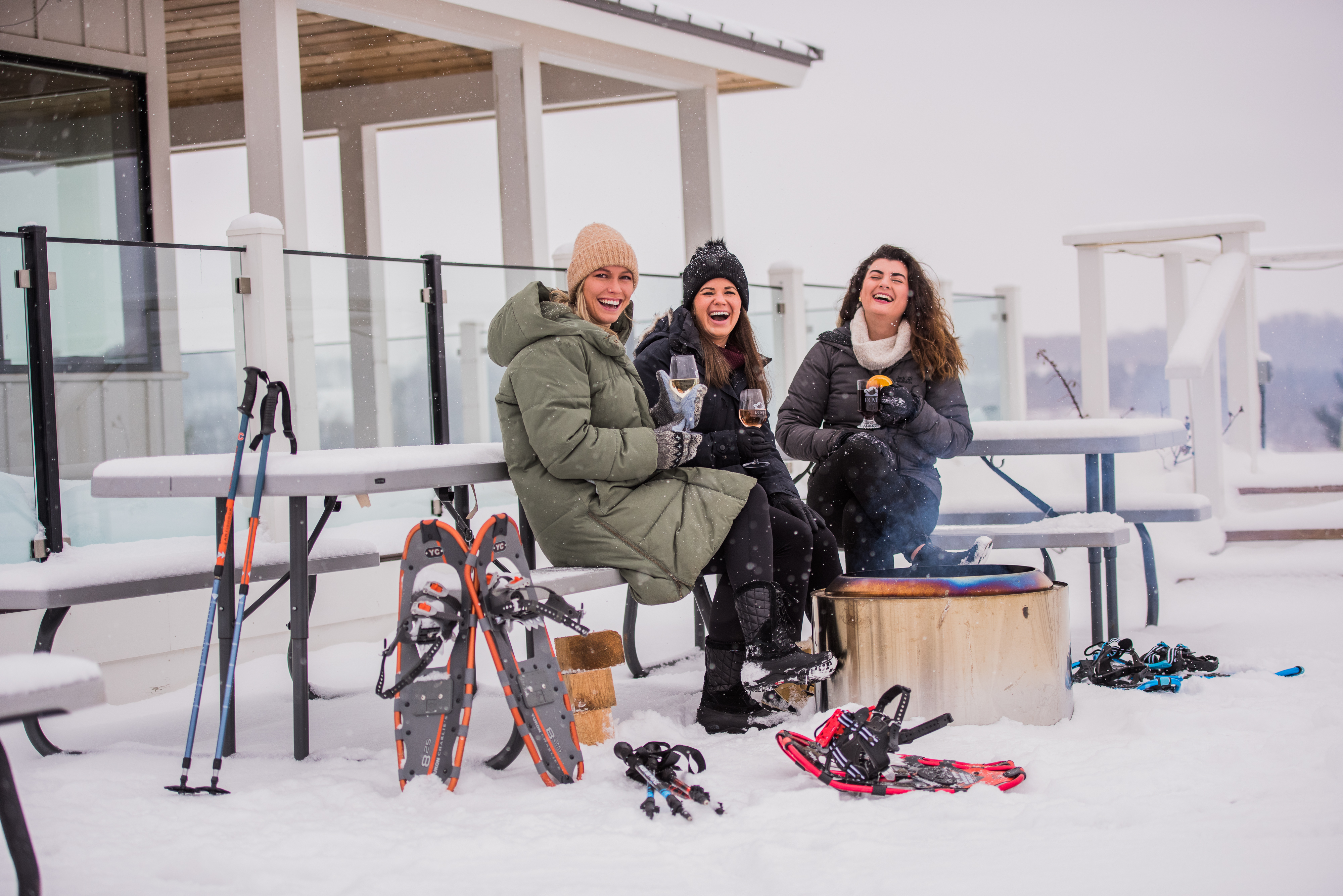 A group of friends sit outside on a snowy day with snowshoes and glasses of wine