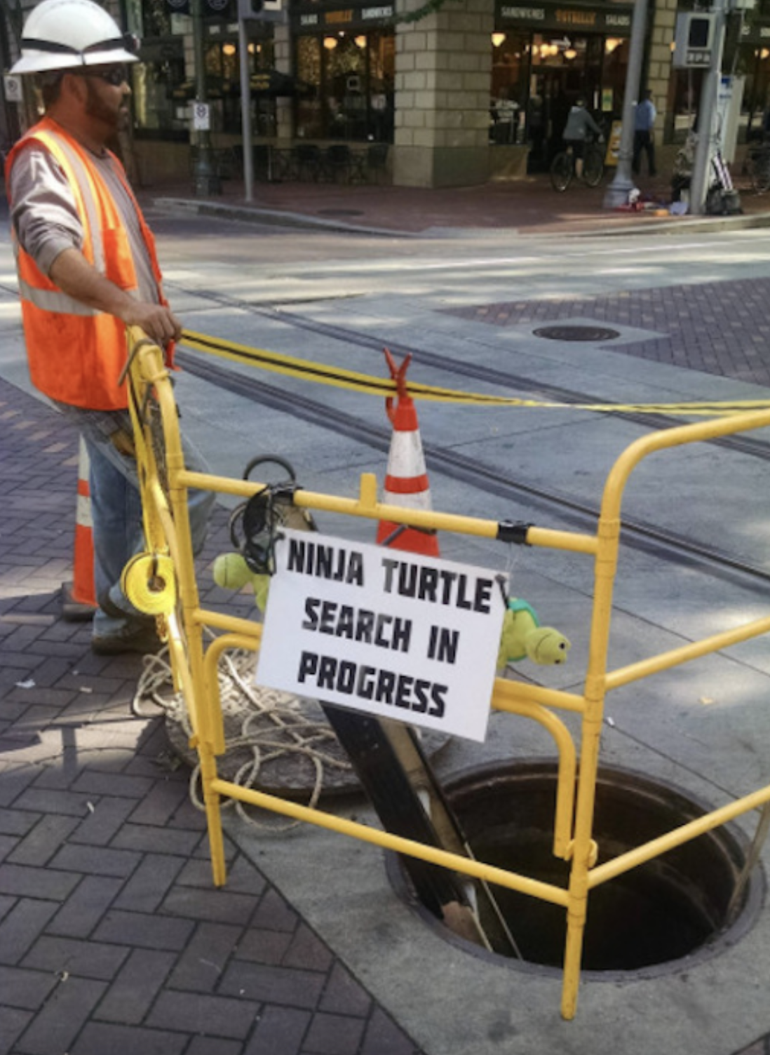 construction worker guarding a sewer hole with sign that reads, ninja turtle search in progress