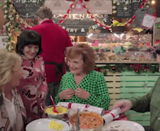 A Christmas-decorated room is full of a long table that pans down of people eating.
