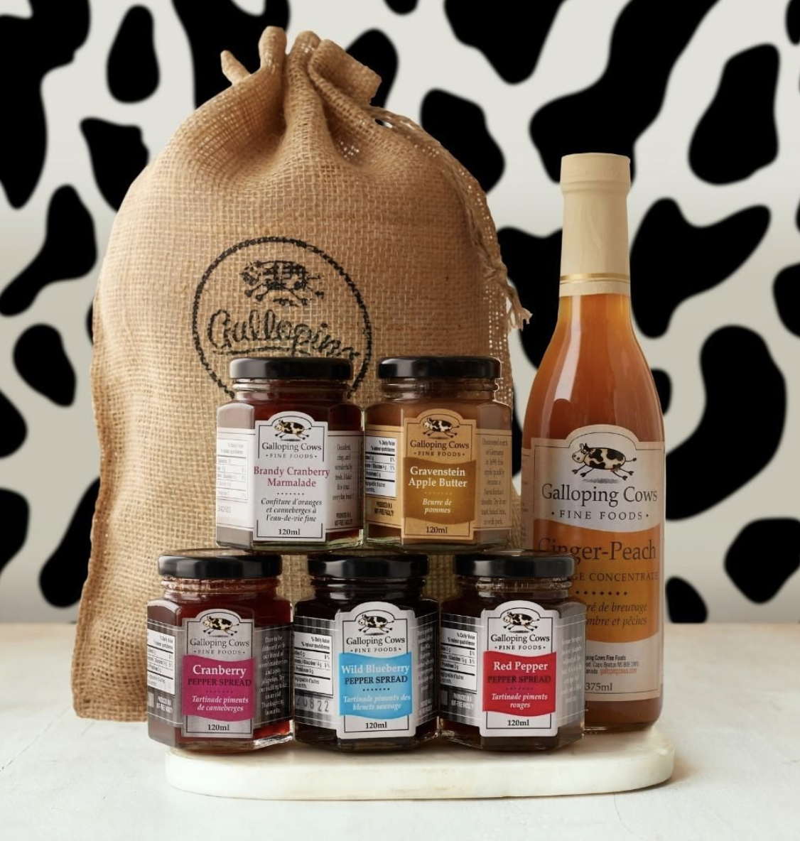 A gift set of different jams and ginger peach bottle, in front of a burlap bag.